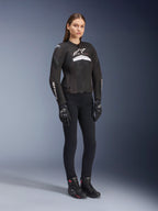 Kvindelig kører iført Alpinestars Stella T-GP Plus R V4 Airflow-jakke, sort og hvid, med mesh-paneler til ventilation og hvide logoer, kombineret med sorte bukser, sorte handsker og sorte støvler med pink detaljer, sportsligt motorcykeludstyr til kvinder