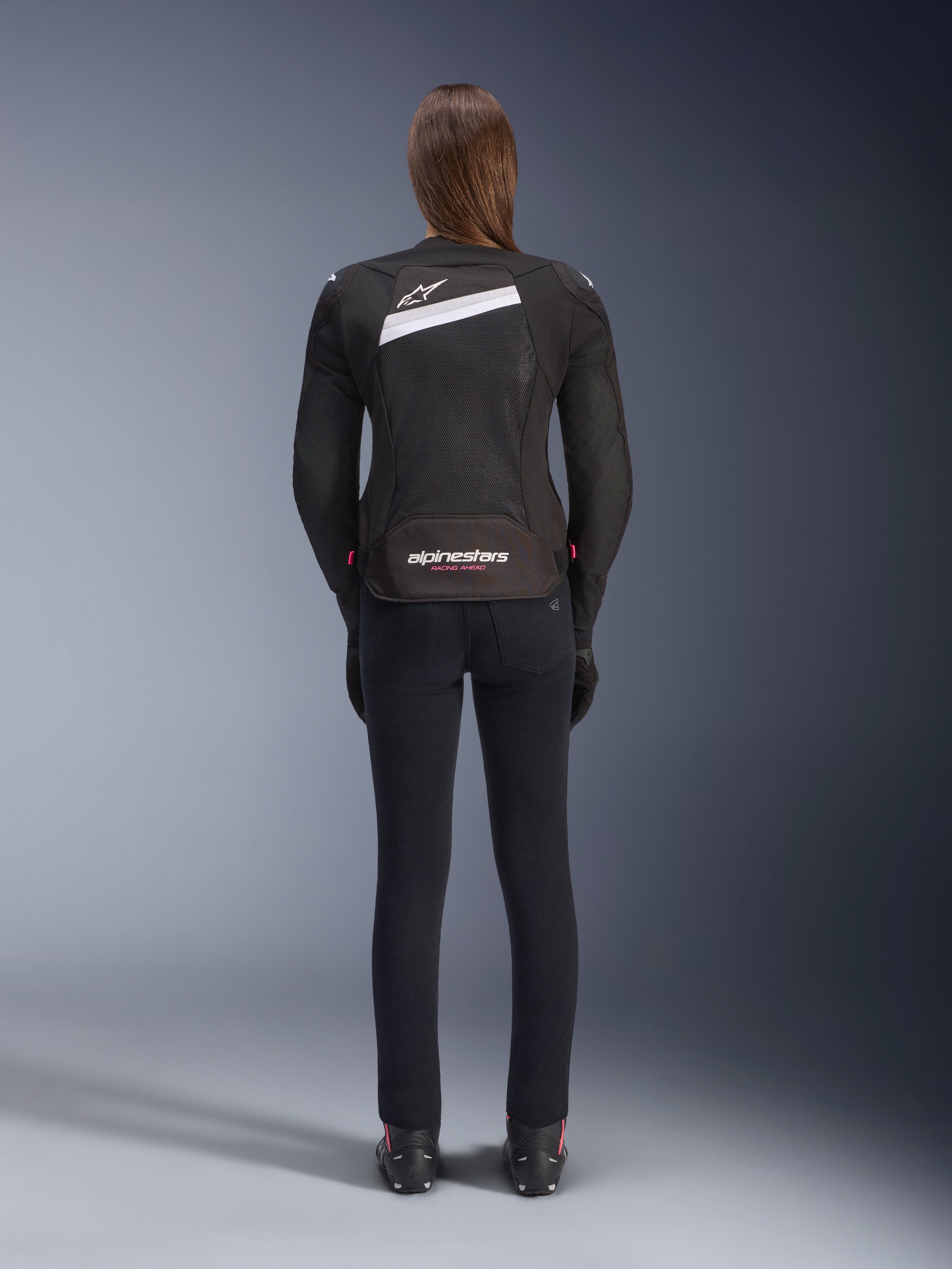 Alpinestars Stella T-GP Plus R V4 Airflow-jakke, sort og hvid, set bagfra med omfattende mesh-ventilationspaneler og Alpinestars-branding, båret af en kvindelig motorcyklist med sorte slim-fit motorcykelbukser og støvler, sport-fit tekstiludstyr til kvinder