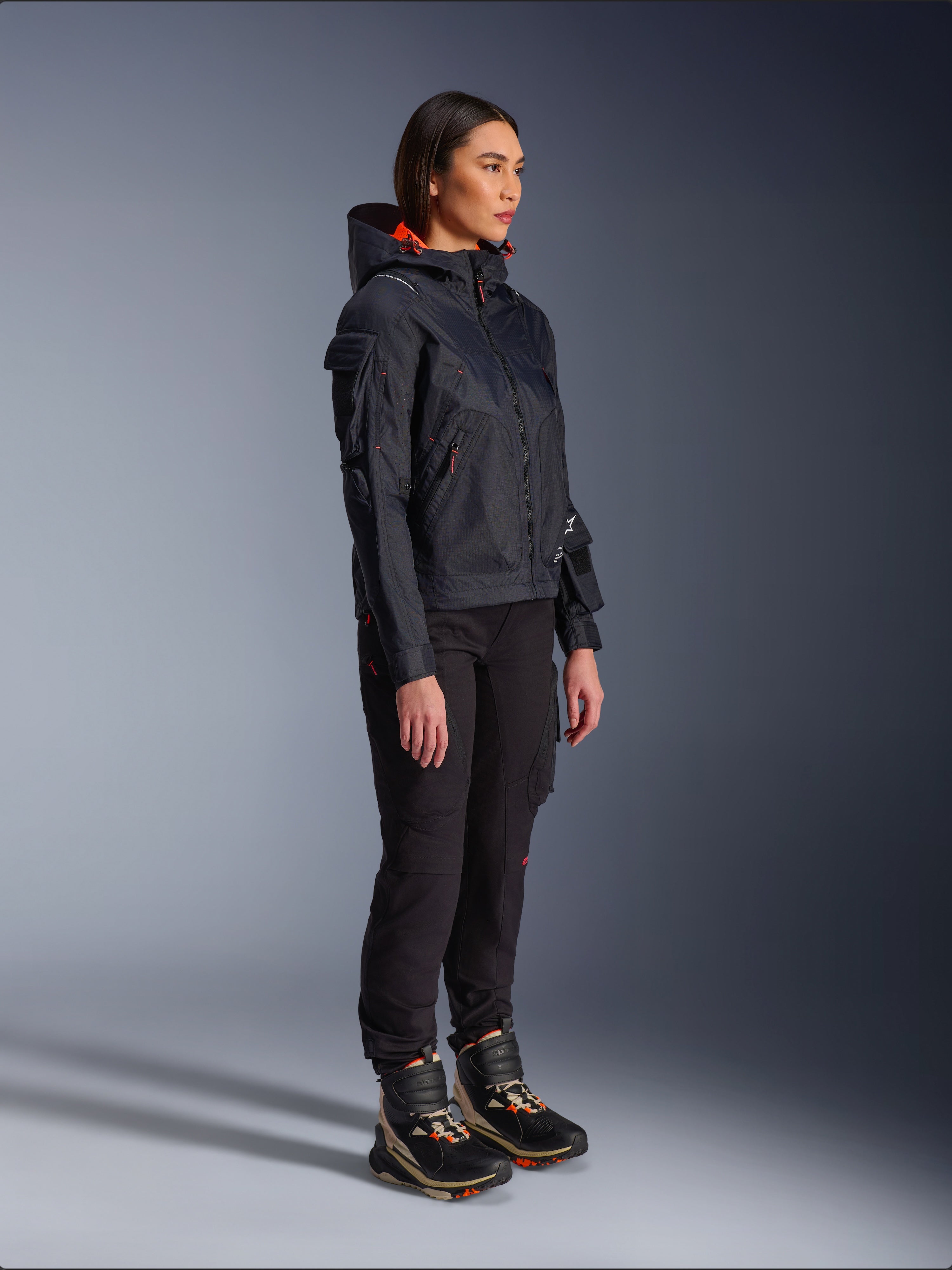 Female rider wearing the Alpinestars Molly Air Jacket in Black Fluo Coral, lightweight textile motorcycle jacket with a bright orange hood lining, styled with black riding pants and technical boots, urban contemporary utility-inspired design.