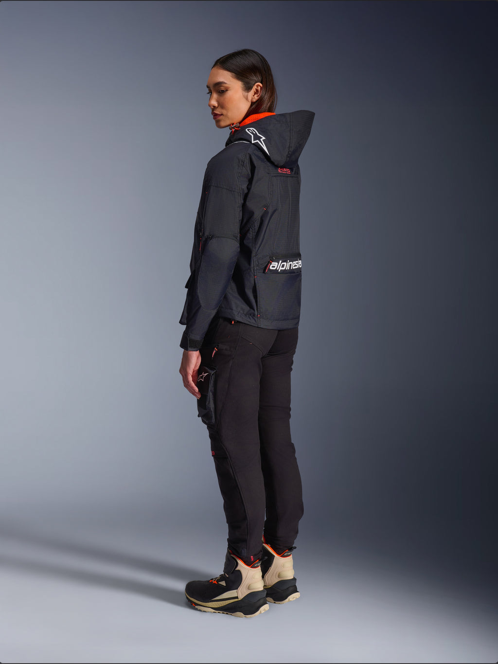 Female rider wearing the Alpinestars Molly Air Jacket in Black Fluo Coral, rear side view showing the hood with Astars logo and large Alpinestars branding on the back, paired with black cargo riding pants and tan and black adventure boots, contemporary urban motorcycle gear for women