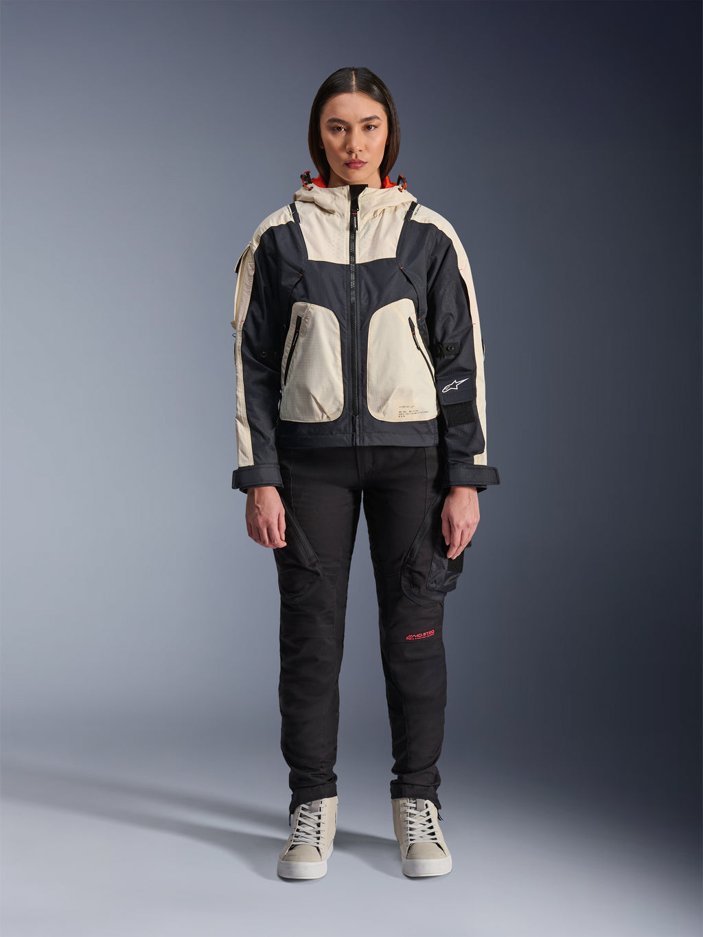 Kvindelig model iført Alpinestars Molly Air-jakke i Black Almond, med et farveopdelt design med cremehvide og sorte paneler og en hætte med rødt for, kombineret med sorte motorcykelbukser og cremehvide sneakers, studieoptagelse af moderne urbant køreudstyr.