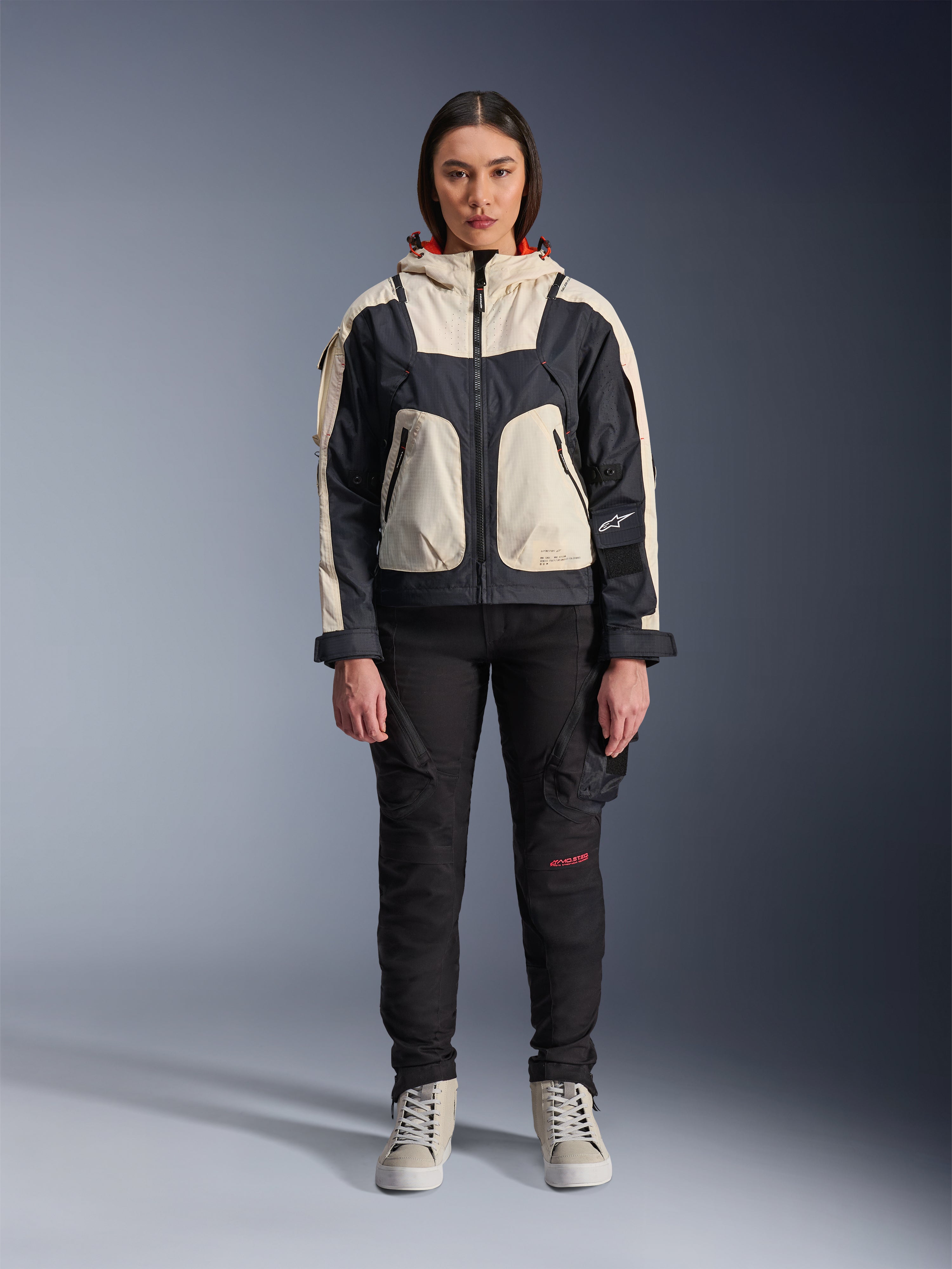 Kvindelig model iført Alpinestars Molly Air-jakke i Black Almond, med et farveopdelt design med cremehvide og sorte paneler og en hætte med rødt for, kombineret med sorte motorcykelbukser og cremehvide sneakers, studieoptagelse af moderne urbant køreudstyr.