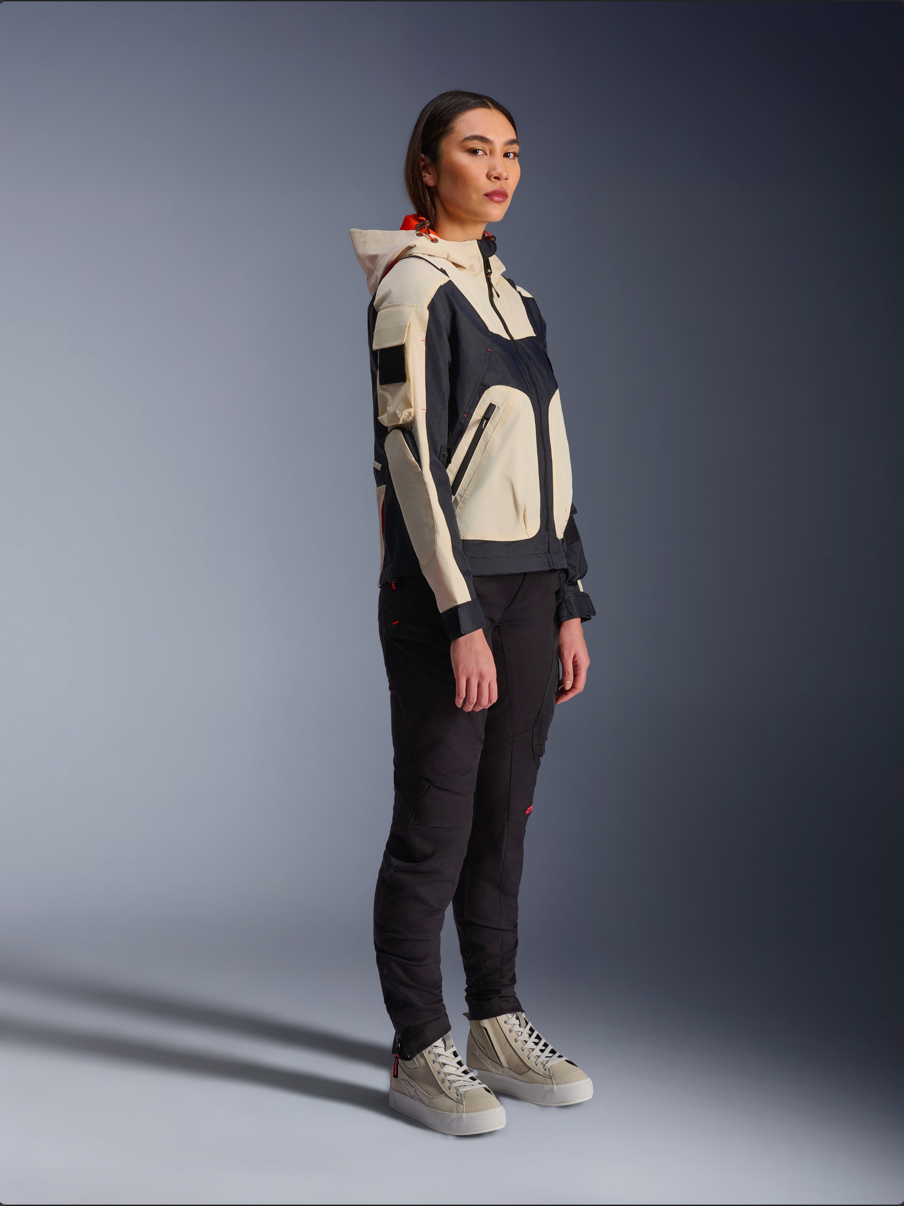 Alpinestars Molly Air-jakke i Black Almond, båret af en kvindelig rytter med sorte bukser og cremefarvede high-top sneakers, totonet design med sorte og beige paneler, integreret hætte og moderne urban stil.