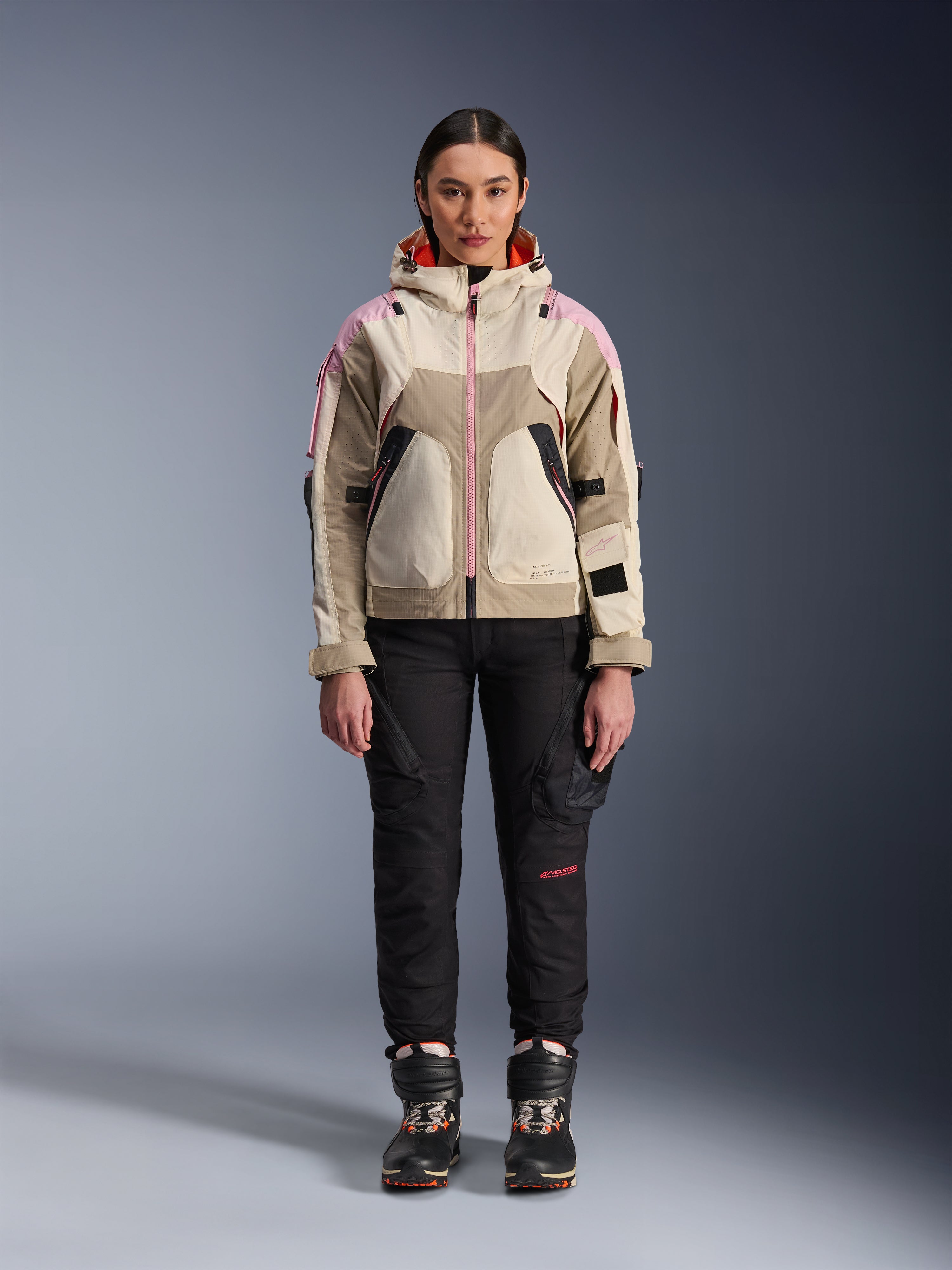 Kvindelig model iført Alpinestars Molly Air-jakke i Sand Pink Almond, kombineret med sorte tekniske kørebukser og støvler, fuldkropsvisning forfra, urban motorcykeljakke med utility-inspireret design og lyserøde detaljer