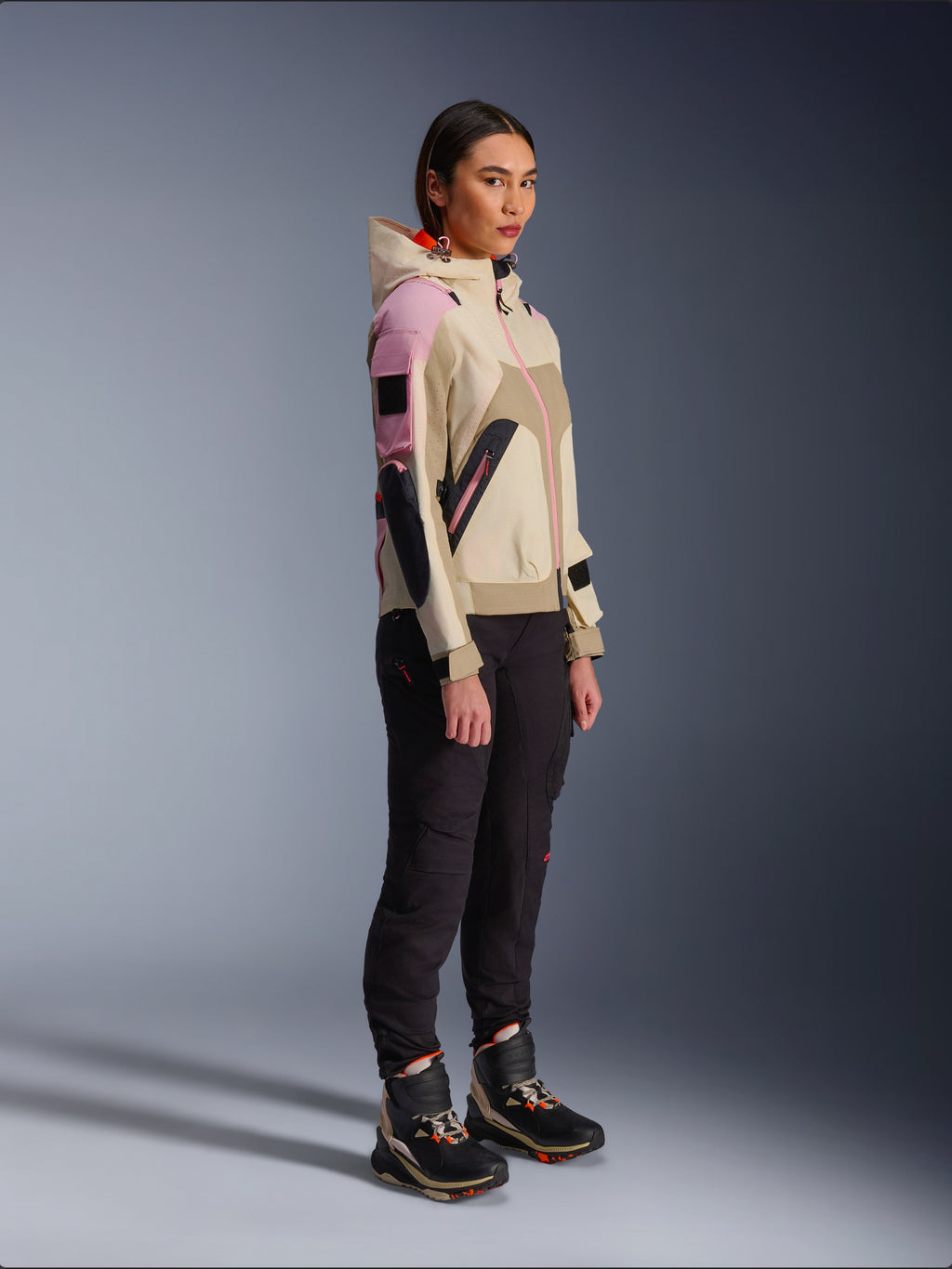Kvindelig model iført Alpinestars Molly Air-jakke i Sand Pink Almond, kombineret med mørke tekniske bukser og high-top kørestøvler, urban motorcykeljakke med hætte og flerfarvede paneler, moderne streetwear-inspireret design