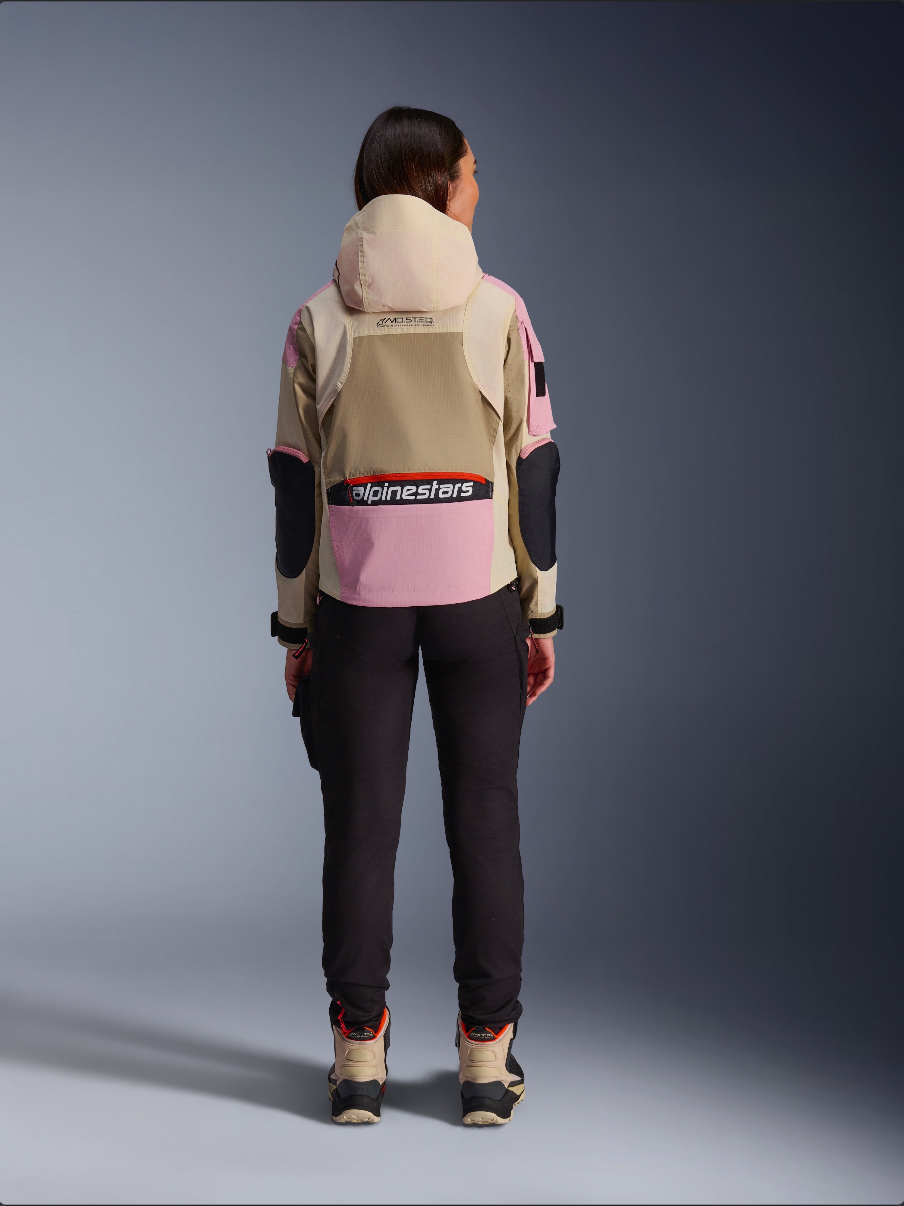 Kvindelig rytter set bagfra iført Alpinestars Molly Air-jakke i Sand Pink Almond, med beige hætte og lyserøde paneler med sorte detaljer, sorte ridebukser og beige motorcykelstøvler, urbant moderne motorcykelbeklædning