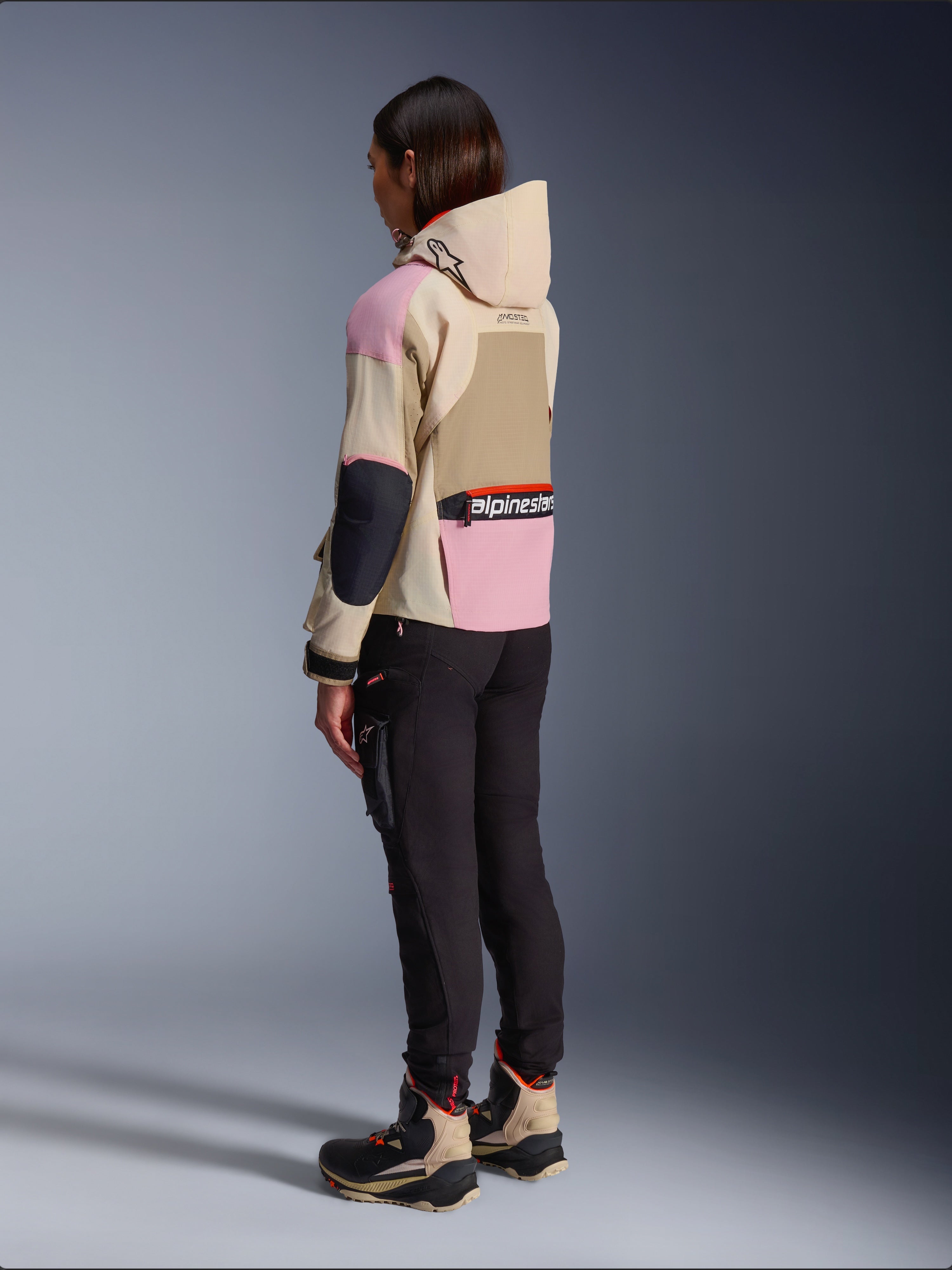 Alpinestars Molly Air Jakke, urban motorcykelstil med hætte, i farven Sand Pink Almond med et flerfarvet beige og lyserødt design, vist fra siden bagfra med sorte kørebukser og matchende tekniske støvler.