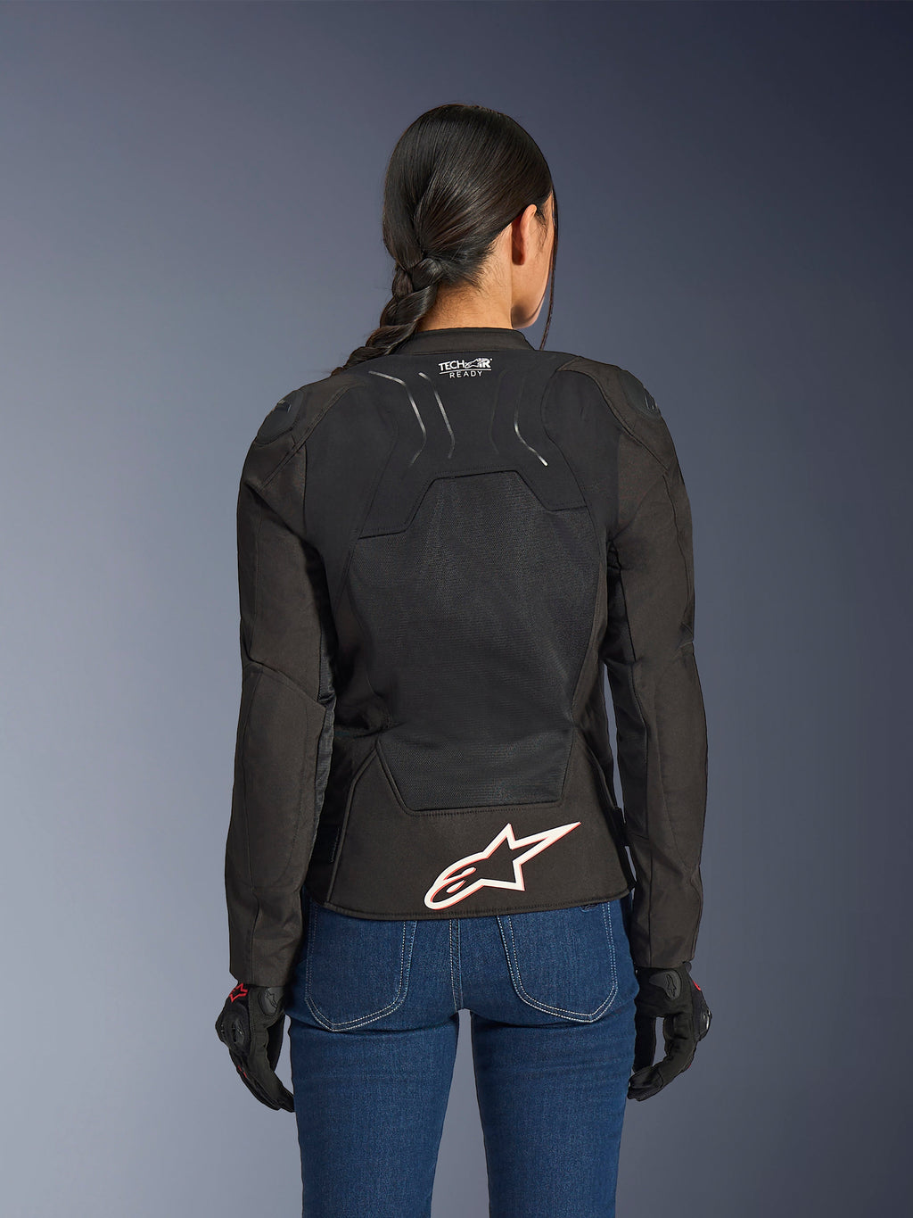 Alpinestars Stella SMX Air-jakke, set bagfra på kvindelig model, sort tekstil med high-flow mesh-paneler, 'TECH-AIR READY'-branding på den øvre ryg, stort hvidt Alpinestars-logo med rød kontur på den nederste del af ryggen, kombineret med blå denimjeans og sorte kørehandsker