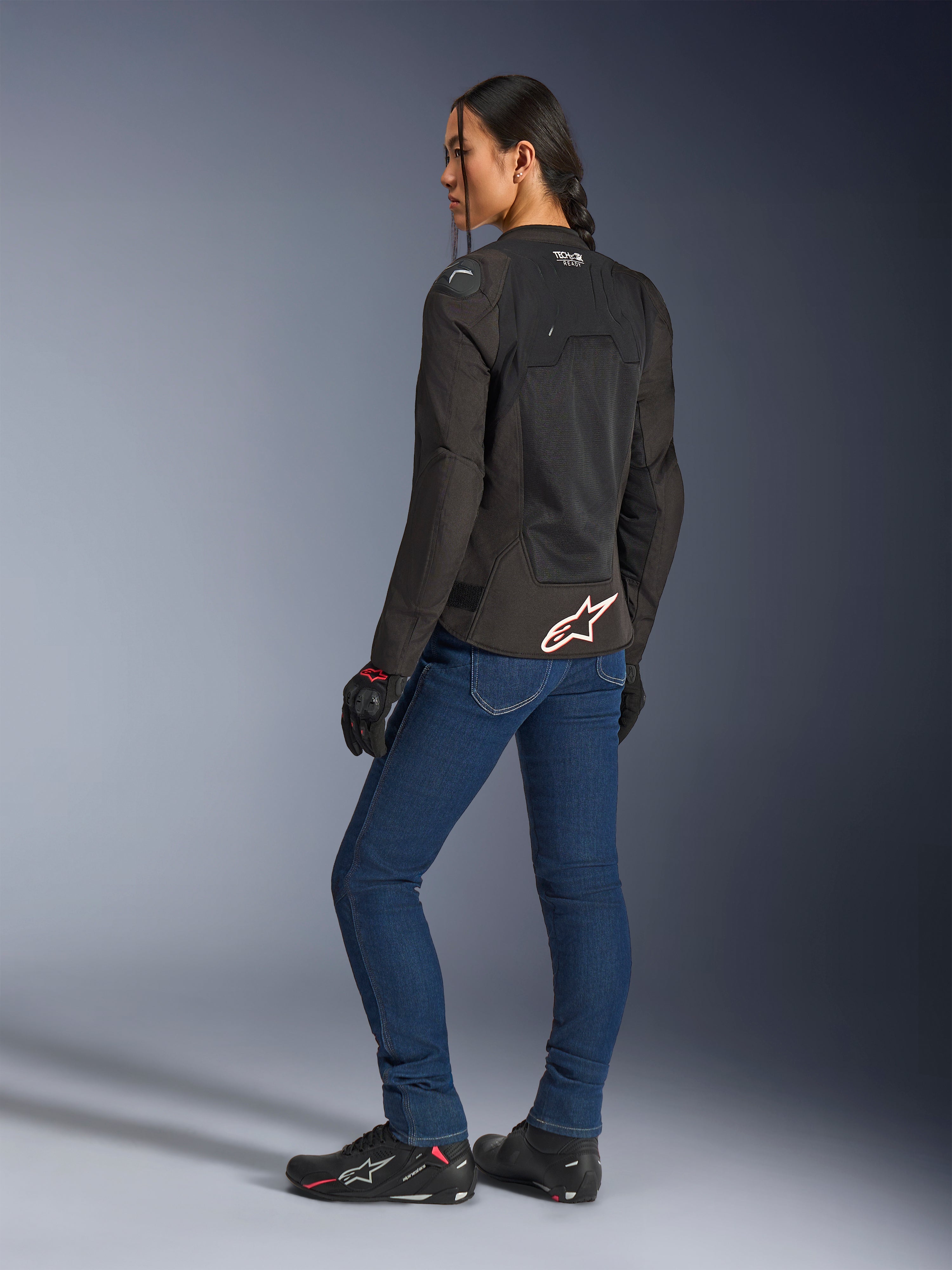 Alpinestars Stella SMX Air jakke, sort med hvidt og rødt logo, båret af en kvindelig kører set skråt bagfra, stylet med blå motorcykeljeans og sort kørefodtøj, teknisk mesh-konstruktion til sportskørsel i varmt vejr