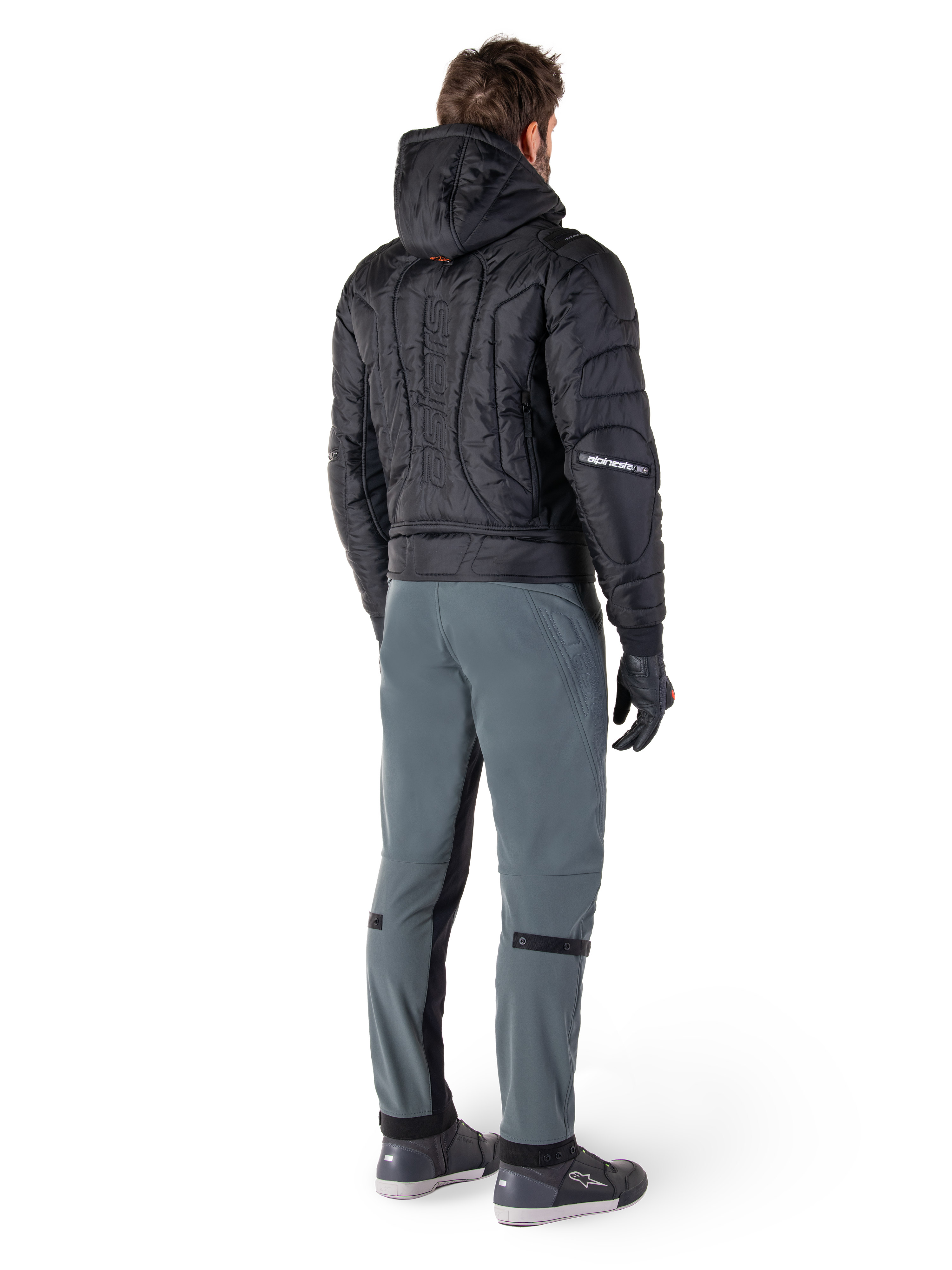 Alpinestars Alden bukser, urbane motorcykelbukser, Urban Gray, set bagfra af en kører iført en sort quiltet jakke med hætte og køre-sneakers, har en slank pasform under knæet med sorte stretch-paneler og knæjusteringer, moderne streetwear-inspireret design.