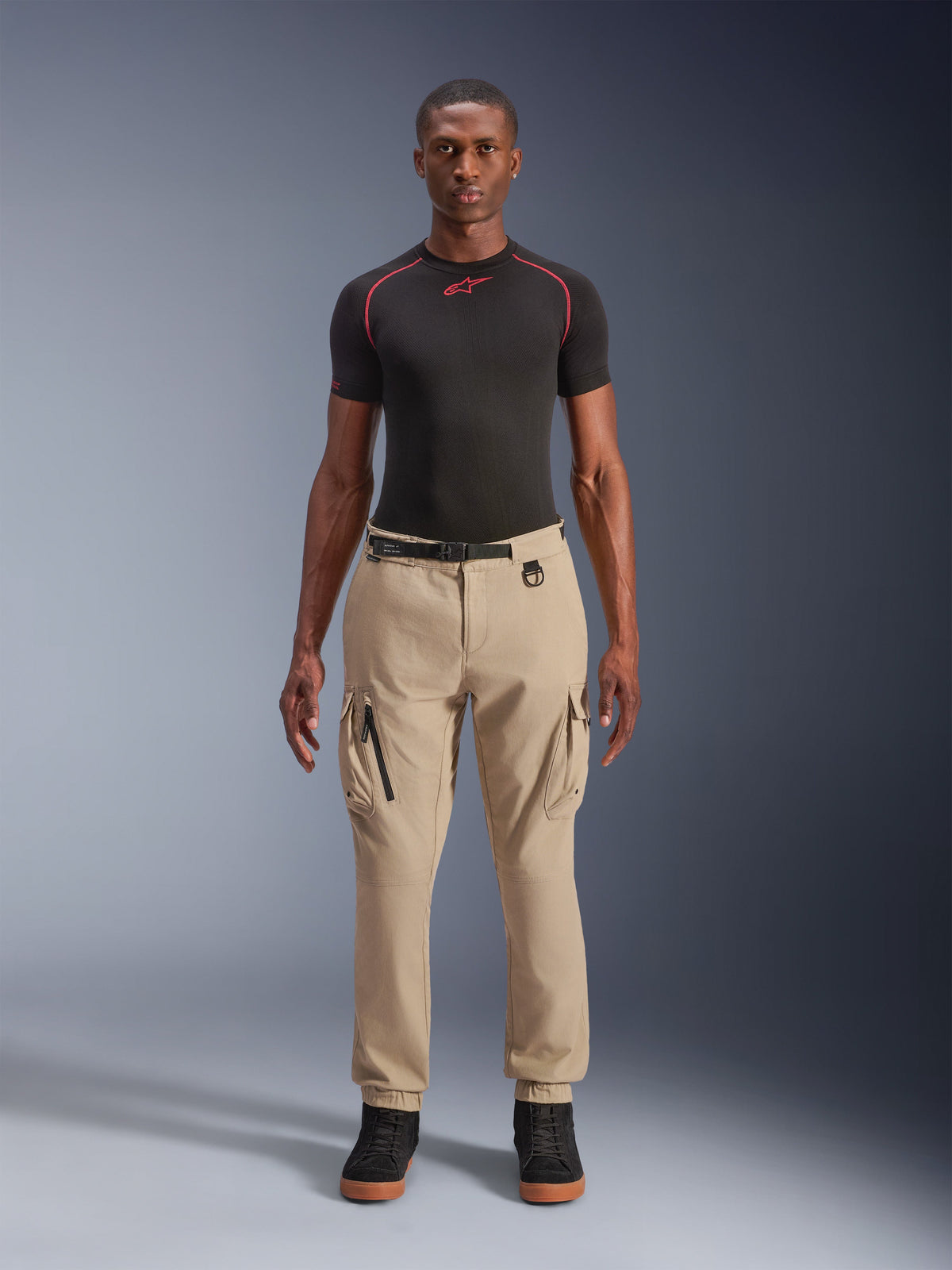Alpinestars Flex-AST Cargo Pants - Short, lys beige urbane motorcykelbukser, regular tapered fit med elastiske sømme, funktionelle cargolommer og integreret sort bælte med spænde, båret af en mandlig model med et sort basislag og sneakers.