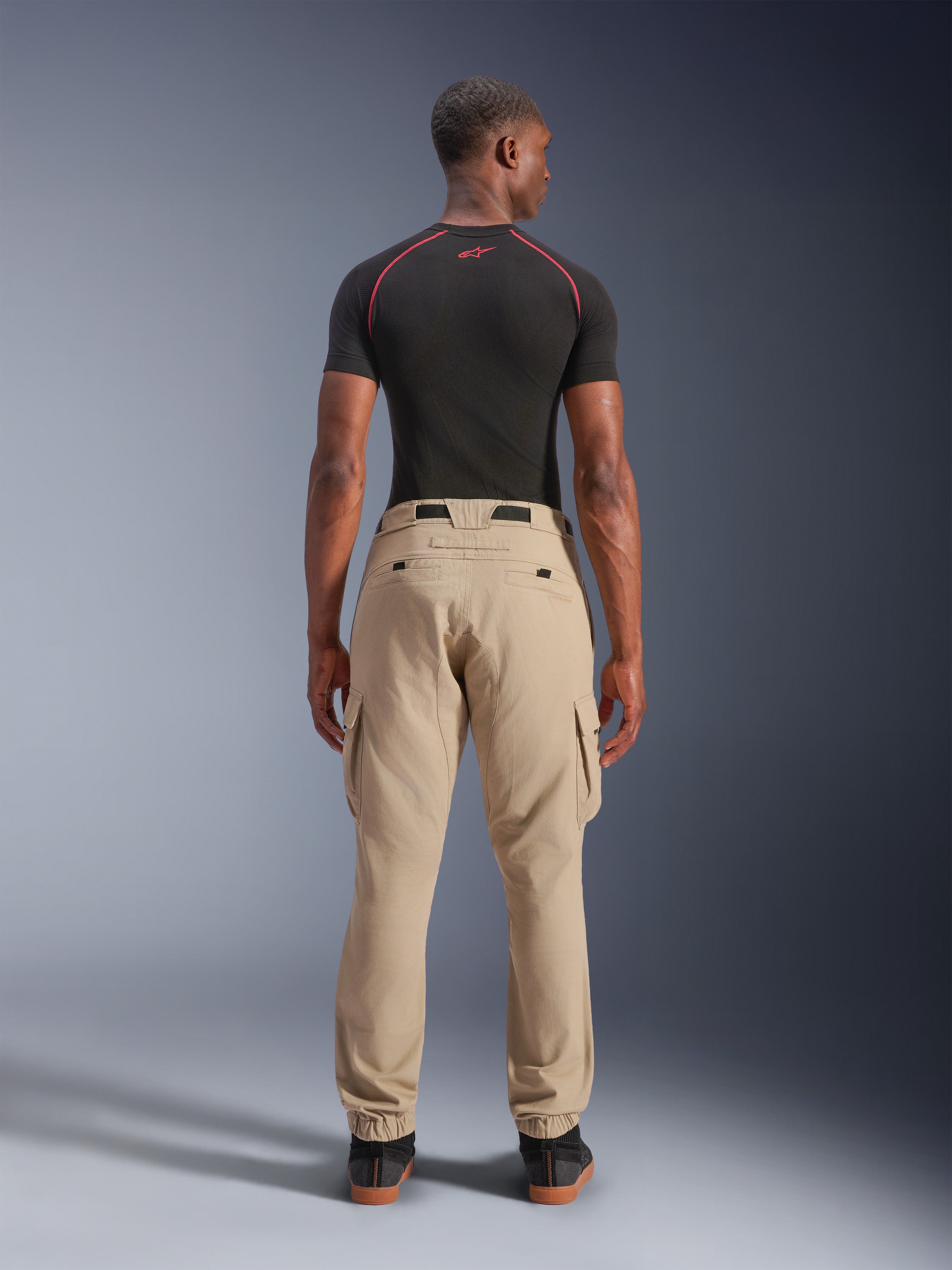 Alpinestars Flex-AST Cargo Pants - Short, lys beige, set bagfra på en model, regular tapered fit med elastiske sømme og cargolommer, båret med et sort teknisk basislag med røde detaljer, moderne urbant motorcykeltøj