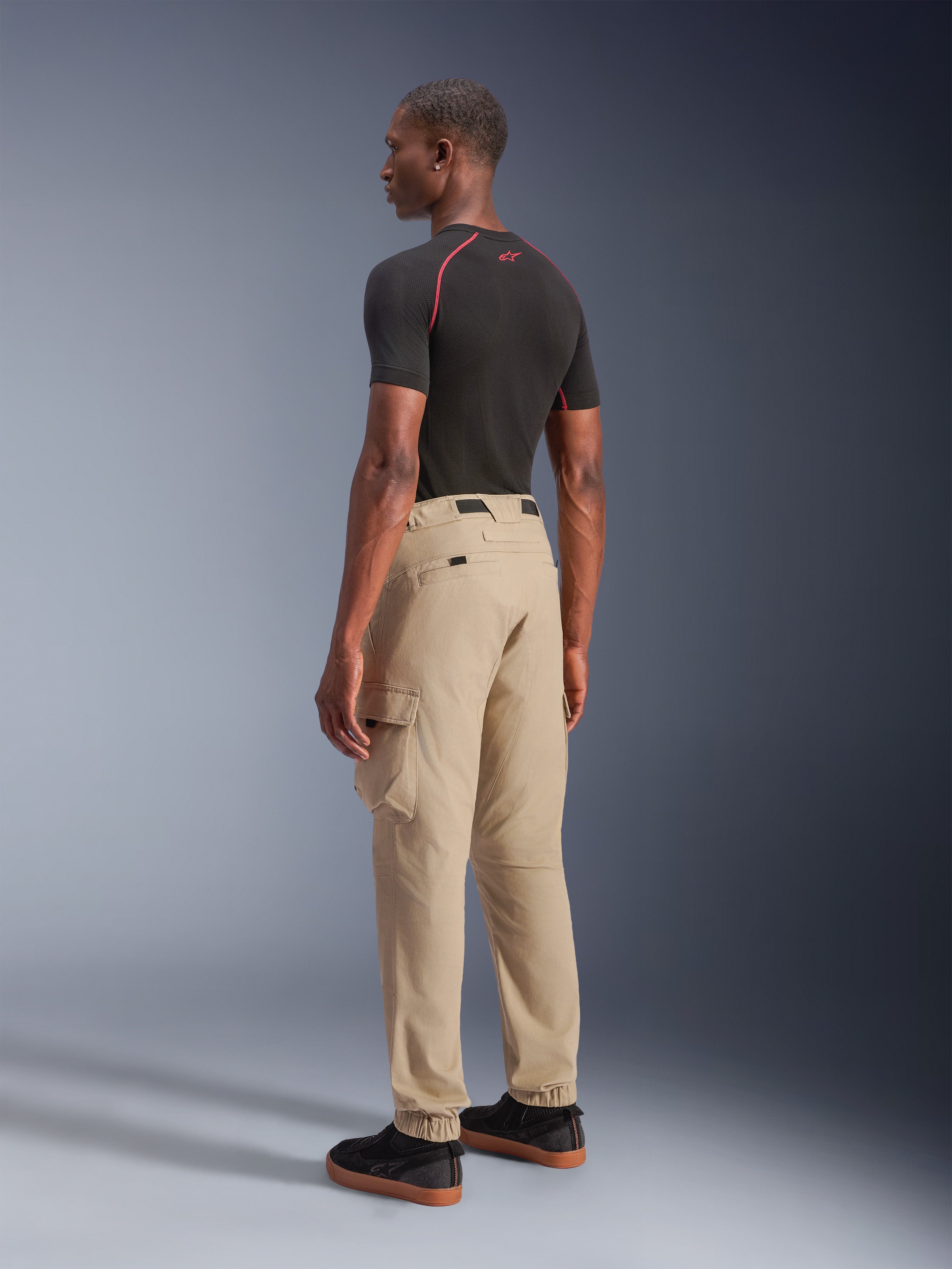 Alpinestars Flex-AST Cargo bukser - Short, urbane motorcykelbukser i lys beige, set bagfra fra siden, viser cargolommer i siden og elastiske opslag, stylet med en sort kortærmet overdel og sorte hverdagssko