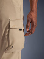 Alpinestars Flex-AST Cargo-bukser, urbane motorcykelbukser, lys beige, nærbillede af cargolomme med tone-i-tone broderet logo, metaløje og sort accentstrop, viser slidstærkt strækbart tekstilmateriale.