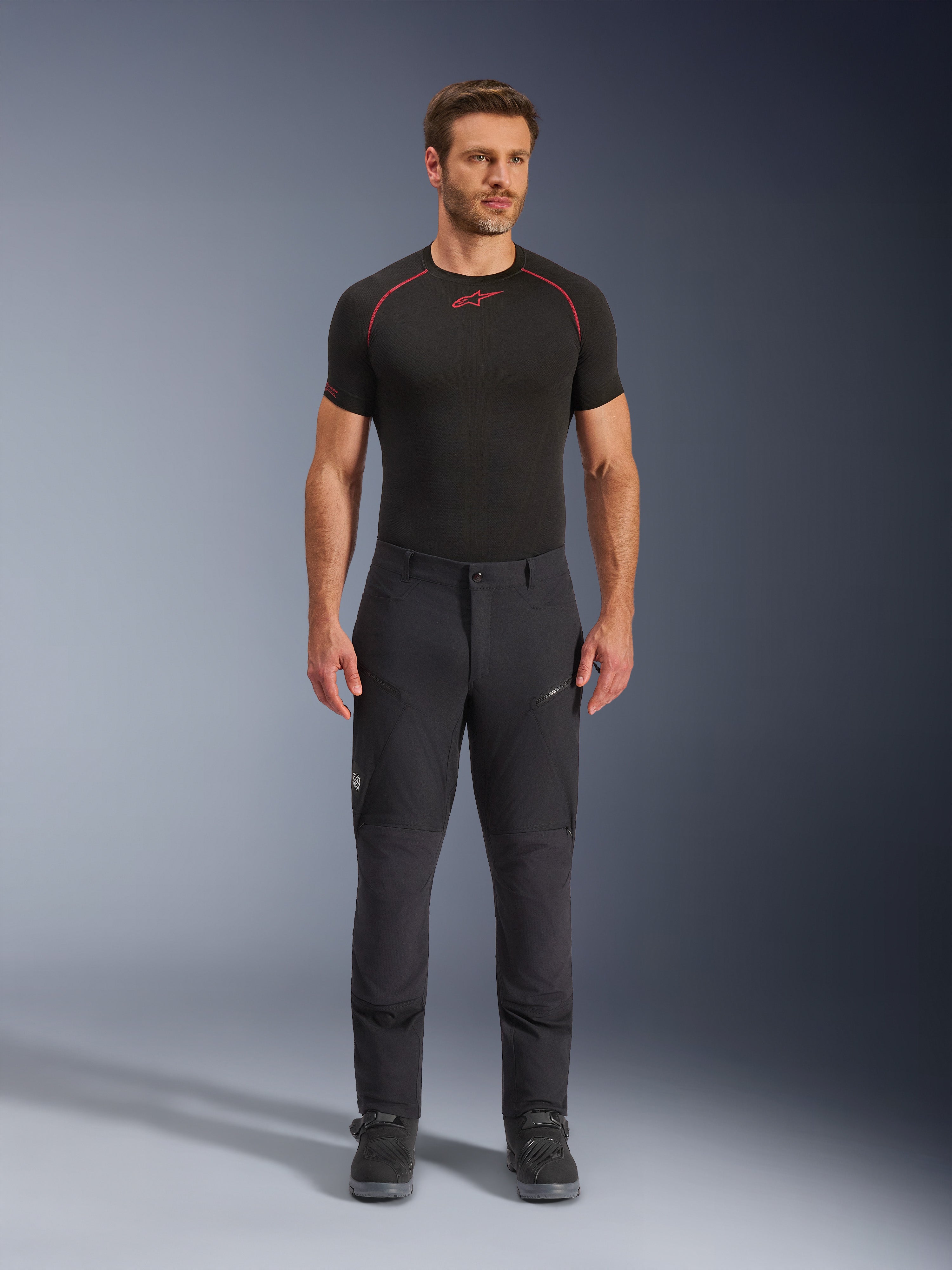 Alpinestars Flex-AST Explorer bukser, sorte adventure touring motorcykelbukser, mand iført tætsiddende tekstilbukser med cargolommer med lynlås og forstærkede paneler, stylet med et sort basislag og motorcykelstøvler, slidstærk beskyttelse til adventure-kørsel