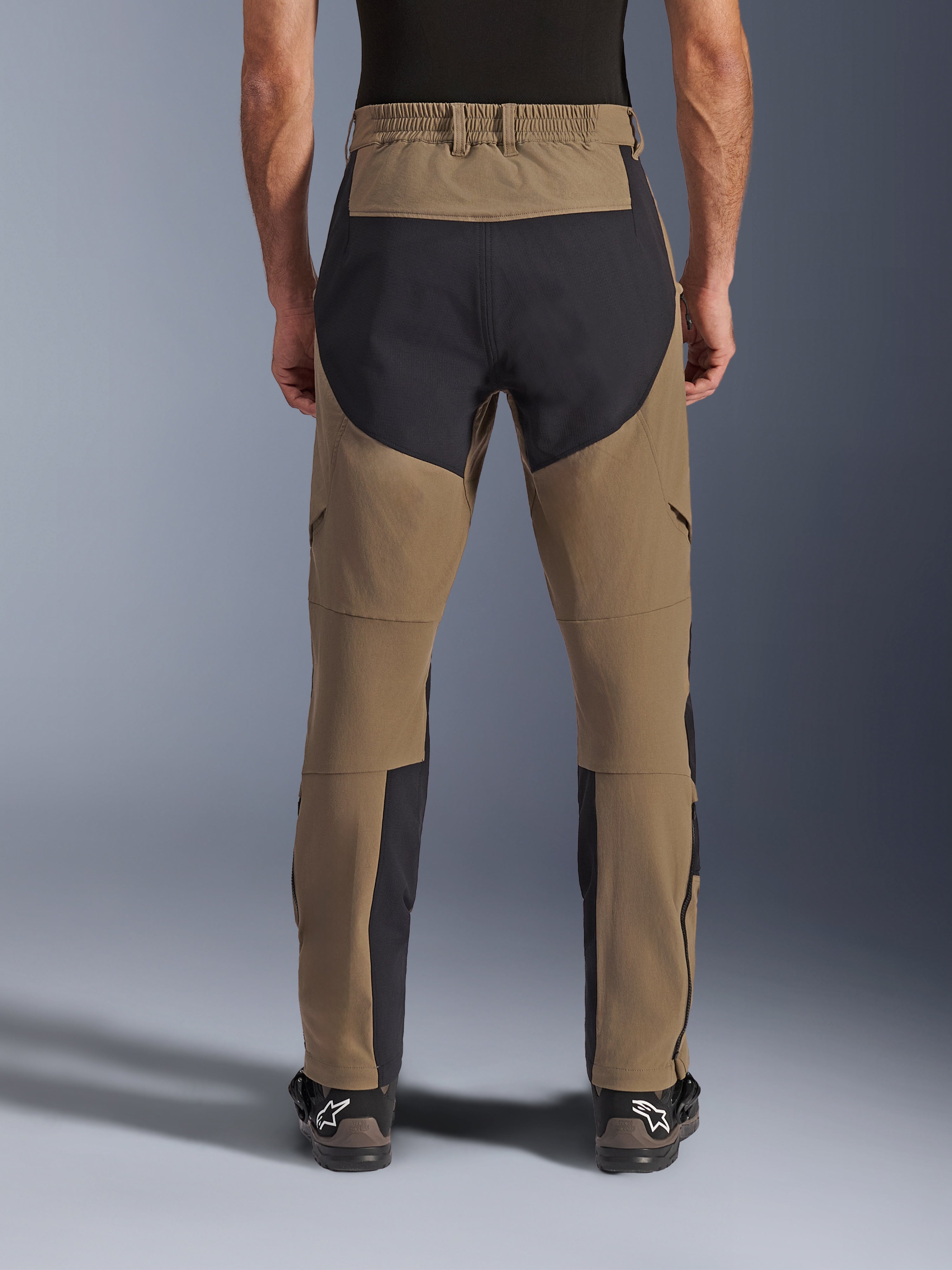Alpinestars Flex-AST Explorer bukser, adventure motorcykelbukser, Walnut Black, set bagfra båret af kører, sorte ripstop-forstærkningspaneler på sædet og indersiden af benene, elastisk linning, lange lynlåse på underbenene og matchende adventure-støvler
