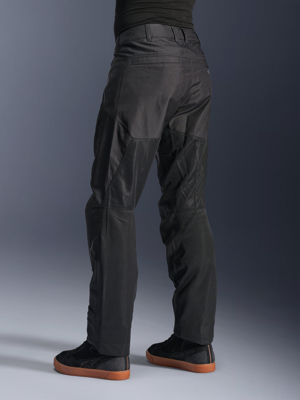 Alpinestars C-1 Air bukser, urbane motorcykelbukser, sorte, set bagfra med store tech mesh-paneler på bagsiden af benene til ventilation, stilrent design til bykørsel, båret med afslappede sorte sneakers.