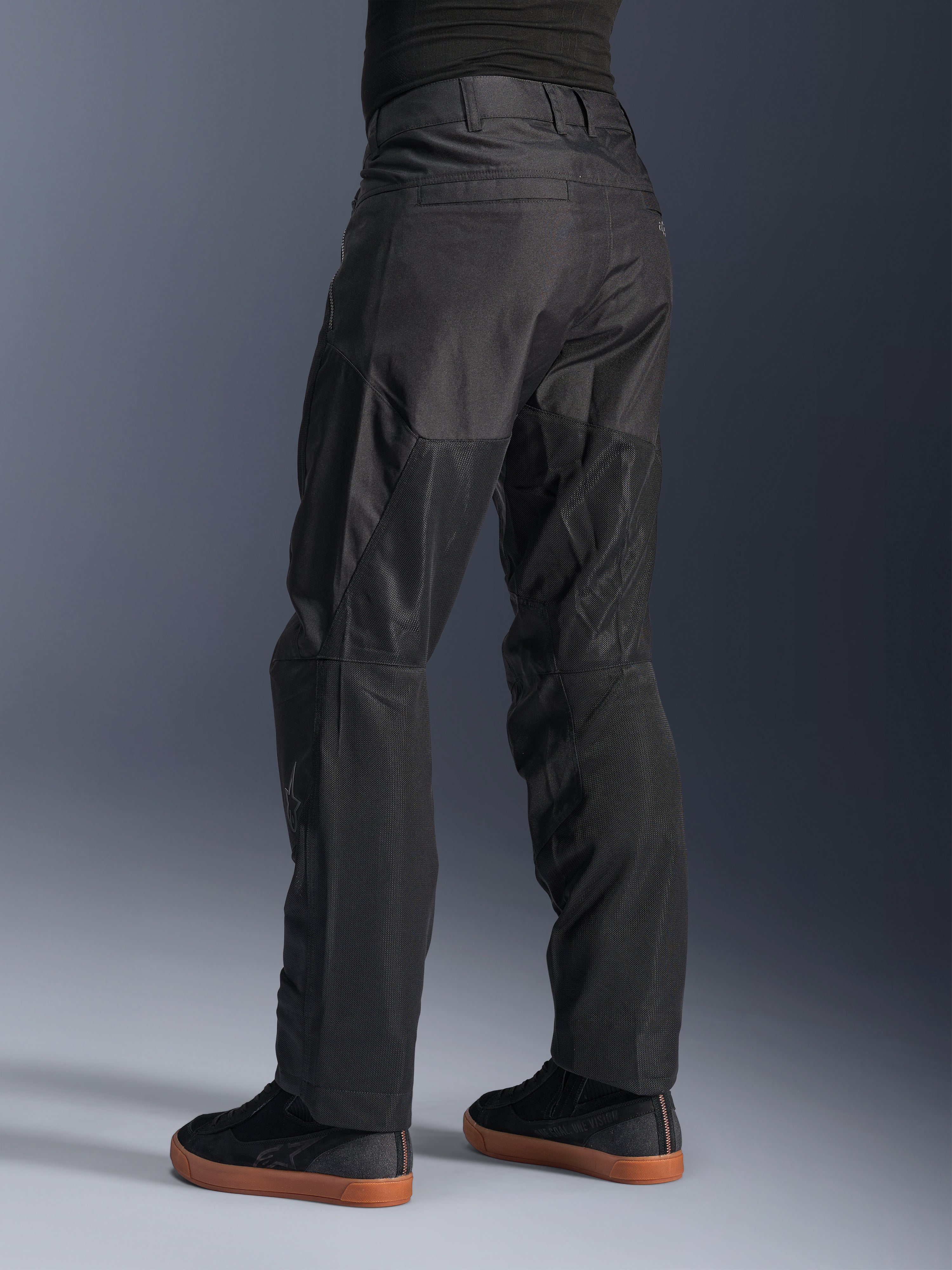 Alpinestars C-1 Air bukser, urbane motorcykelbukser, sorte, set bagfra med store tech mesh-paneler på bagsiden af benene til ventilation, stilrent design til bykørsel, båret med afslappede sorte sneakers.