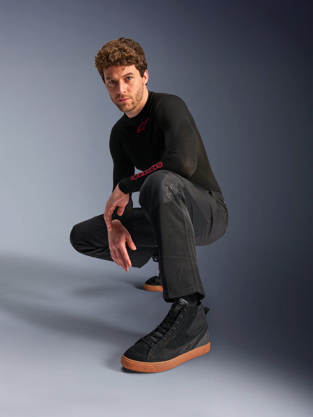 Alpinestars C-1 Air bukser, urban motorcykelbukser, sort, model der sidder på hug i et studiemiljø, båret med en sort langærmet overdel og sorte sneakers med brune såler, med åndbare mesh-paneler og en moderne urban stil