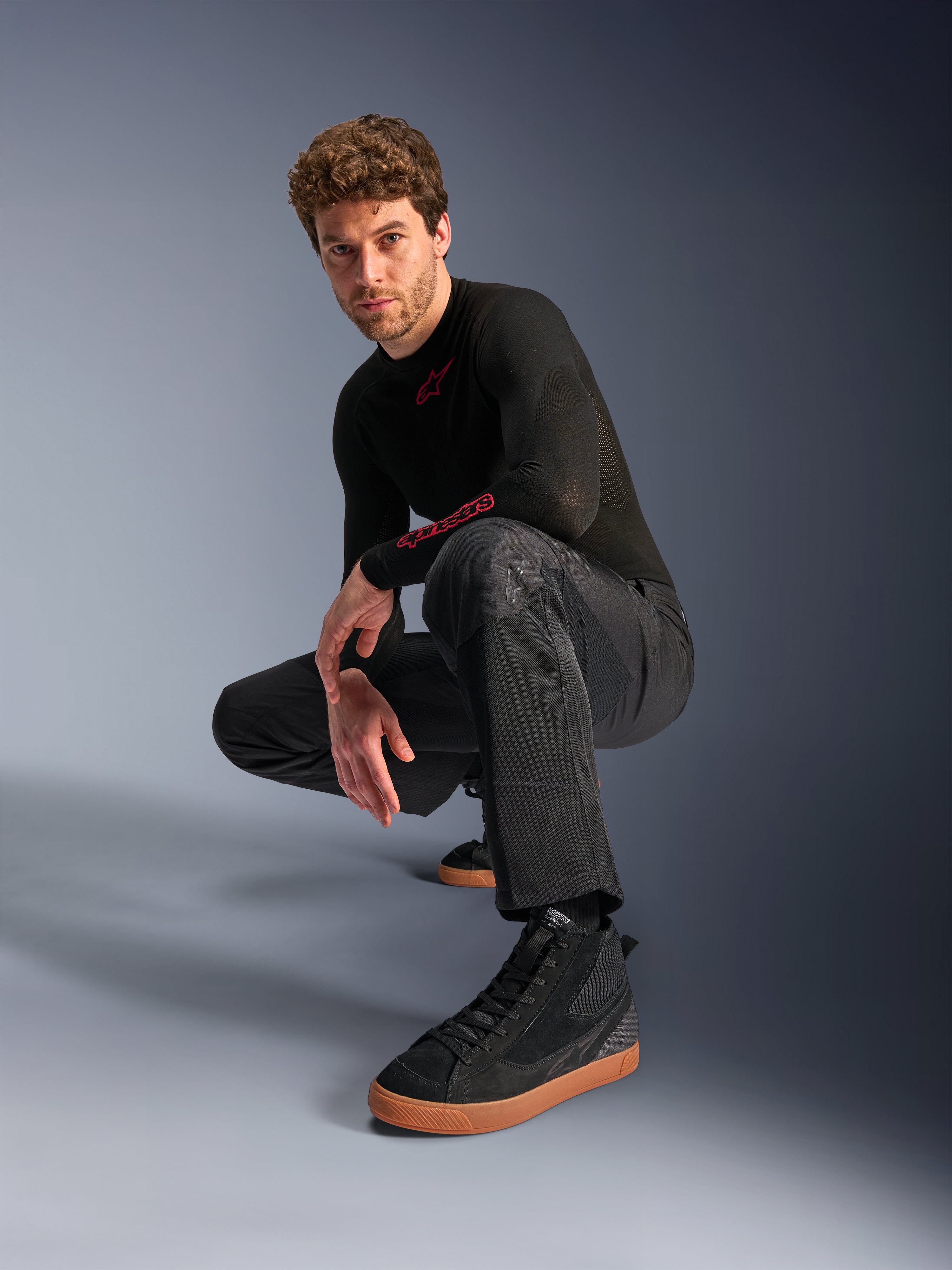Alpinestars C-1 Air bukser, urban motorcykelbukser, sort, model der sidder på hug i et studiemiljø, båret med en sort langærmet overdel og sorte sneakers med brune såler, med åndbare mesh-paneler og en moderne urban stil