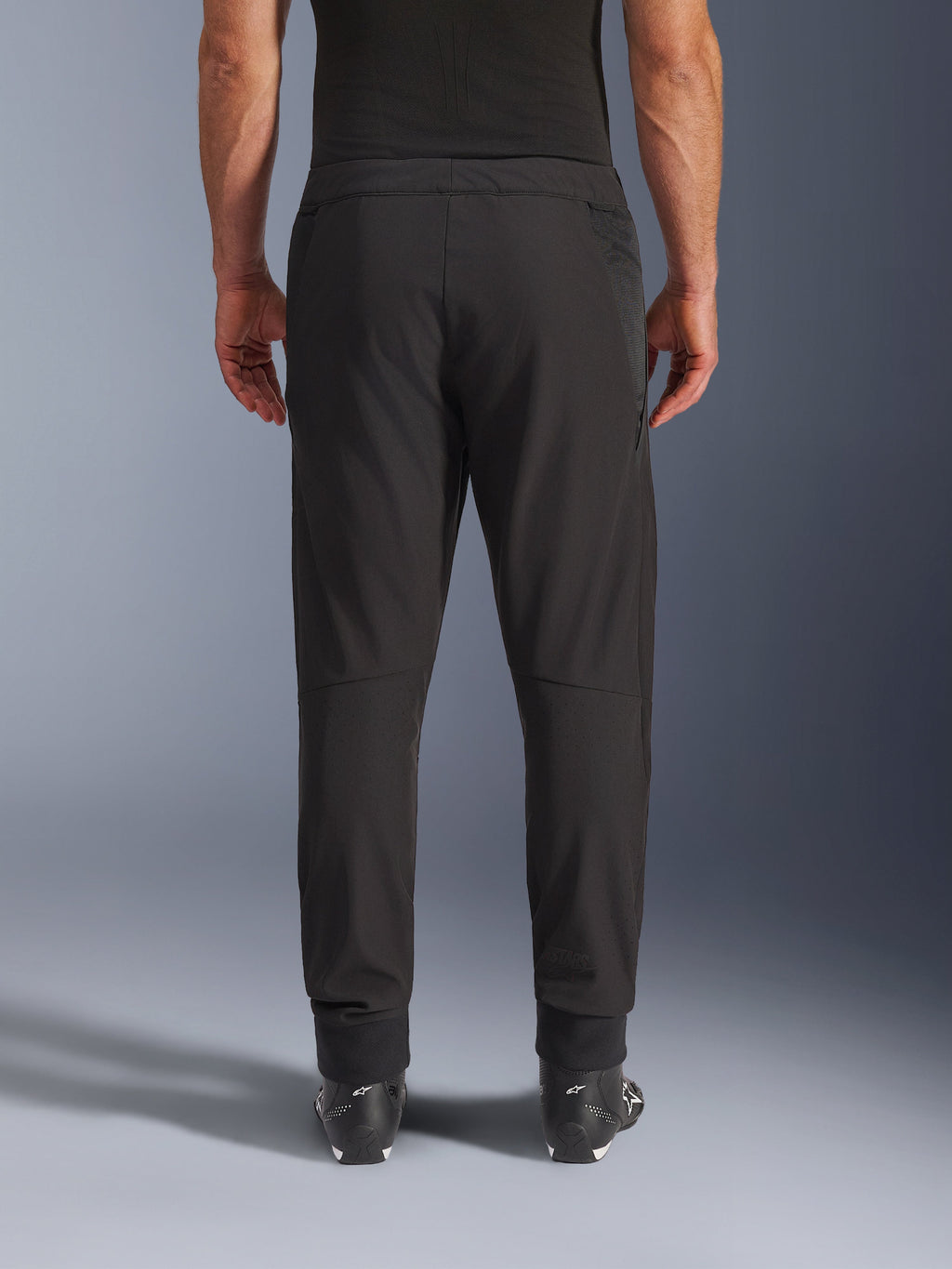 Alpinestars Aeroshell Airflow bukser, sort, set bagfra, med laserperforerede zoner på læggene og mesh-paneler, båret med sorte Alpinestars-støvler, letvægts softshell-design til sport og touring