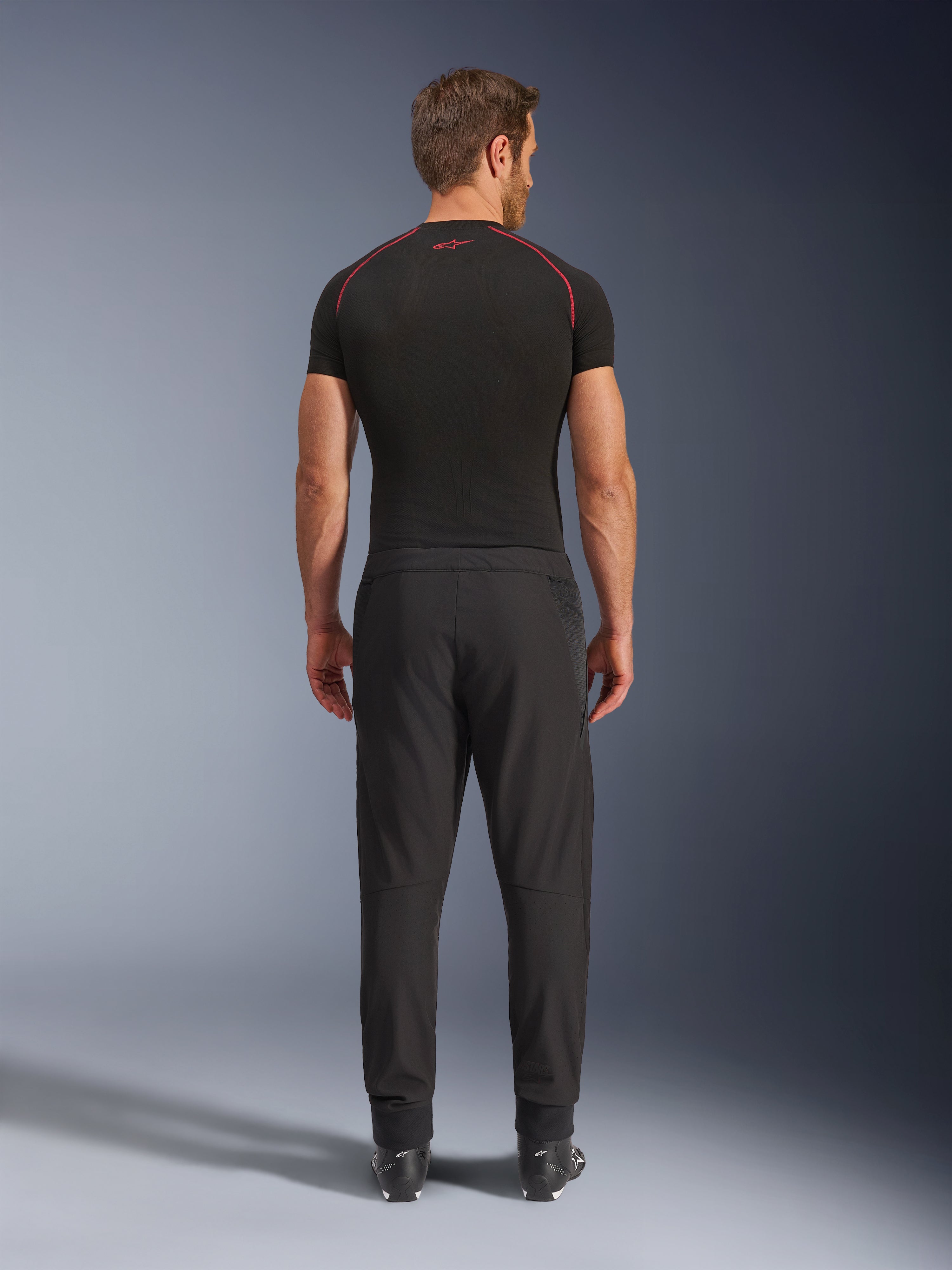 Alpinestars Aeroshell Airflow bukser, sorte motorcykelbukser, set bagfra på en model med et sort baselag og støvler, letvægts softshell-chassis med integrerede mesh-paneler til ventilation
