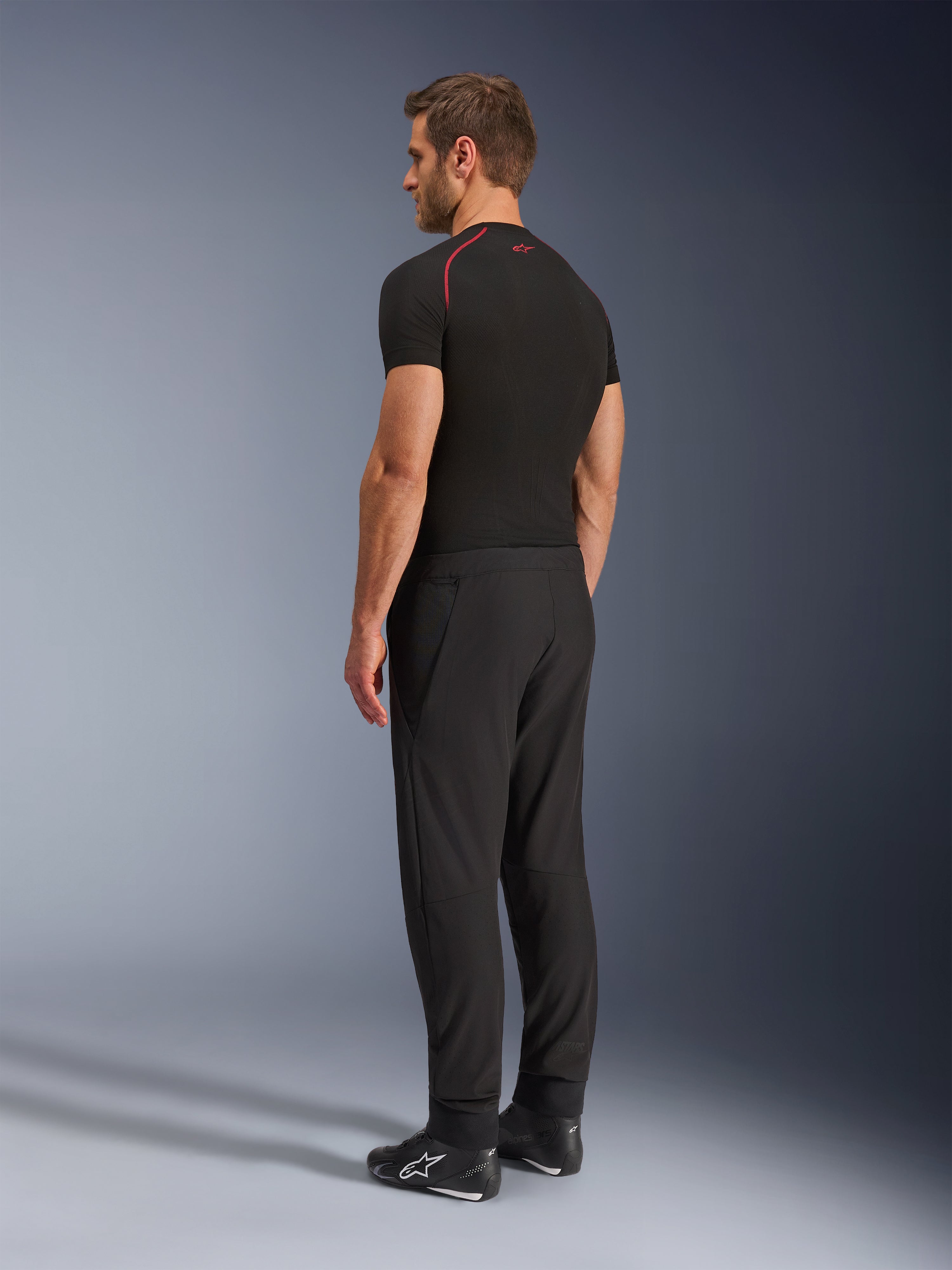 Alpinestars Aeroshell Airflow Bukser, sorte motorcykelbukser, letvægts softshell-tekstil, båret med en sort teknisk kortærmet trøje og sorte kørestøvler, profilvisning bagfra, der viser den slanke pasform og ergonomiske konstruktion.