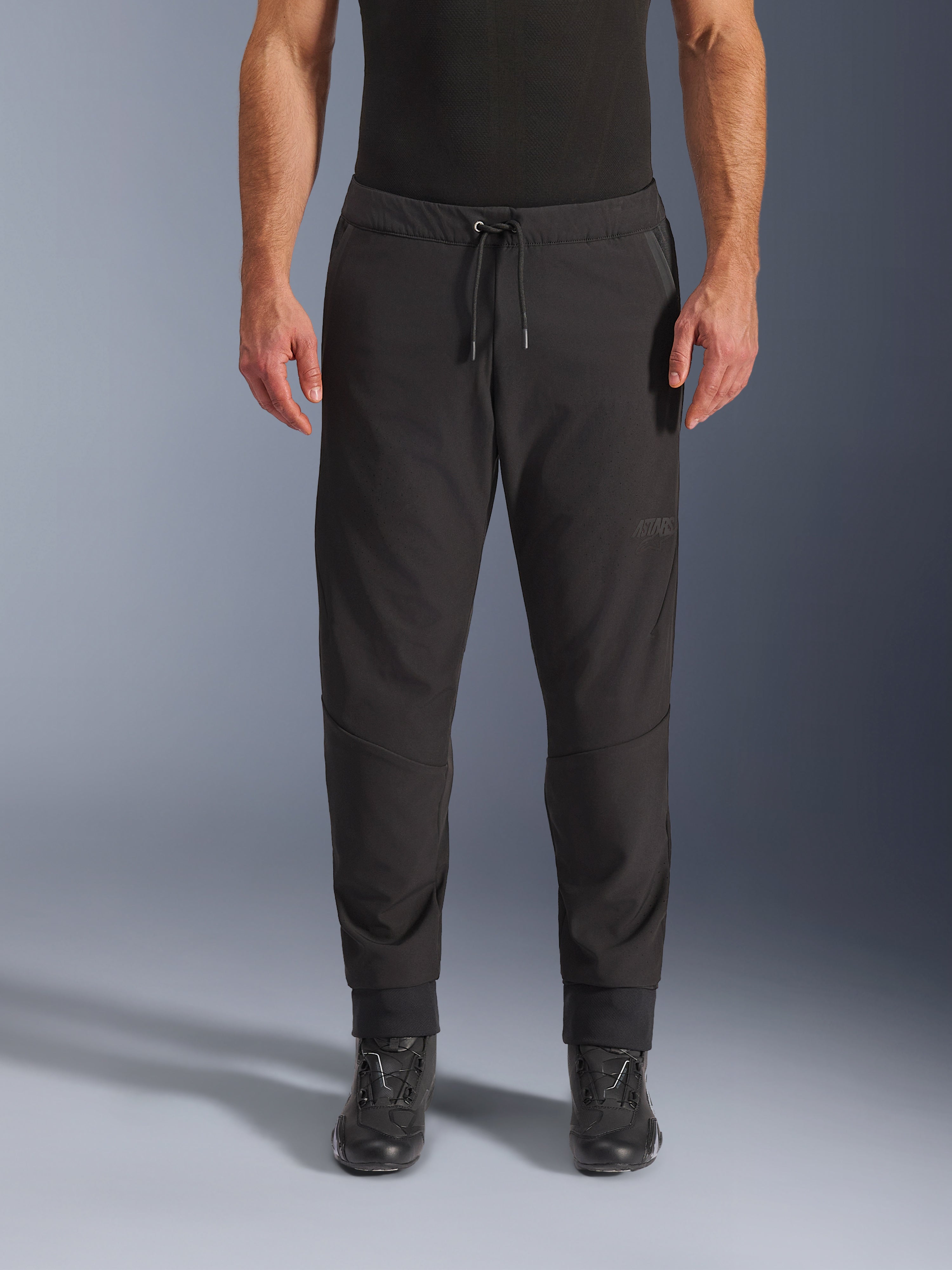 Alpinestars Aeroshell Airflow bukser, sorte motorcykelbukser, set forfra båret med sorte kørestøvler, har snøre i taljen og tætsiddende ankelmanchetter, letvægts softshell-design med diskret tonal branding.