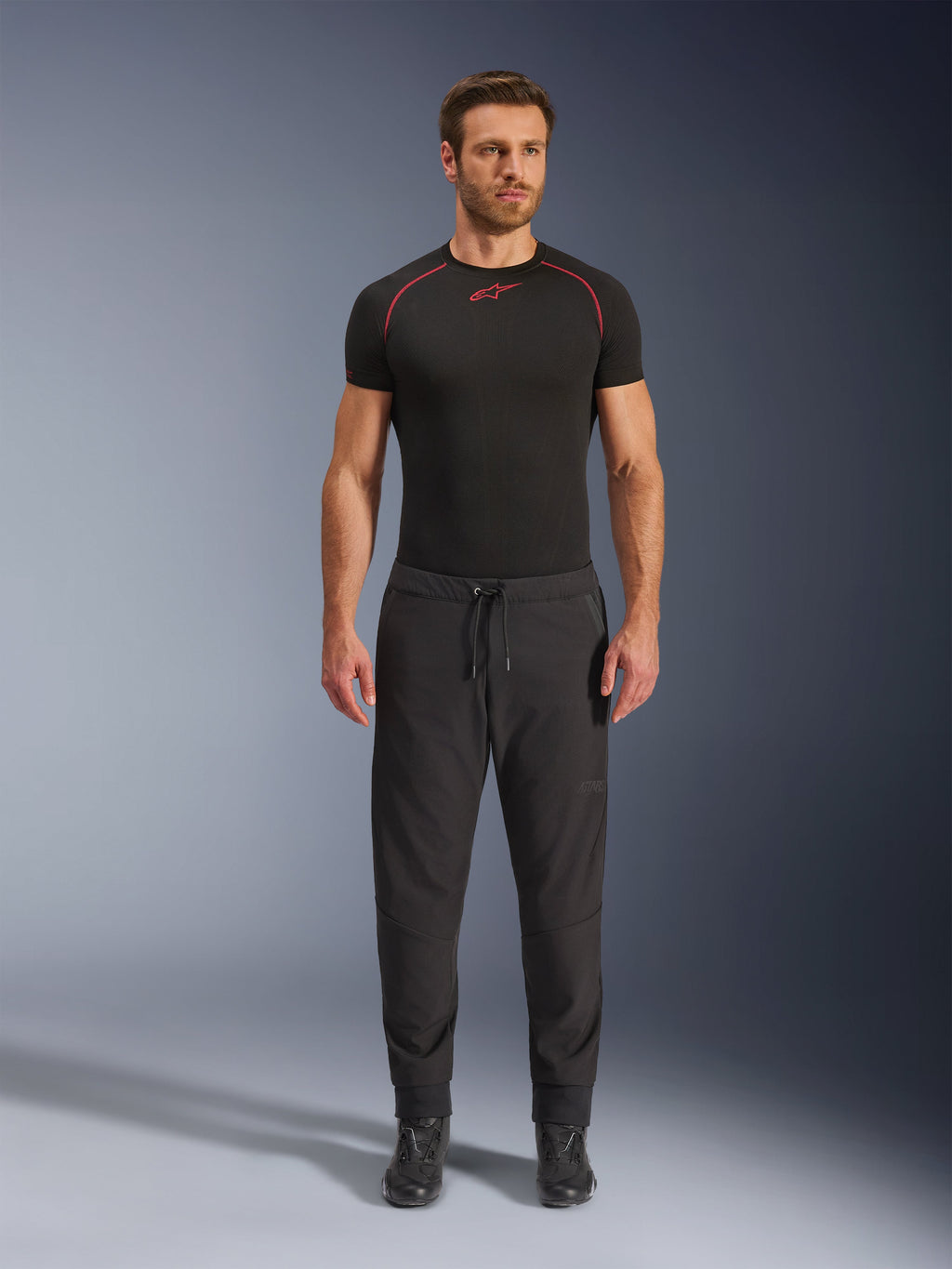 Model iført Alpinestars Aeroshell Airflow bukser i sort, kombineret med et sort kortærmet basislag og sorte motorcykelstøvler. Disse letvægts softshell motorcykelbukser har snoretræk i taljen og elastiske manchetter, designet til sport- og touringkørere under varme vejrforhold.