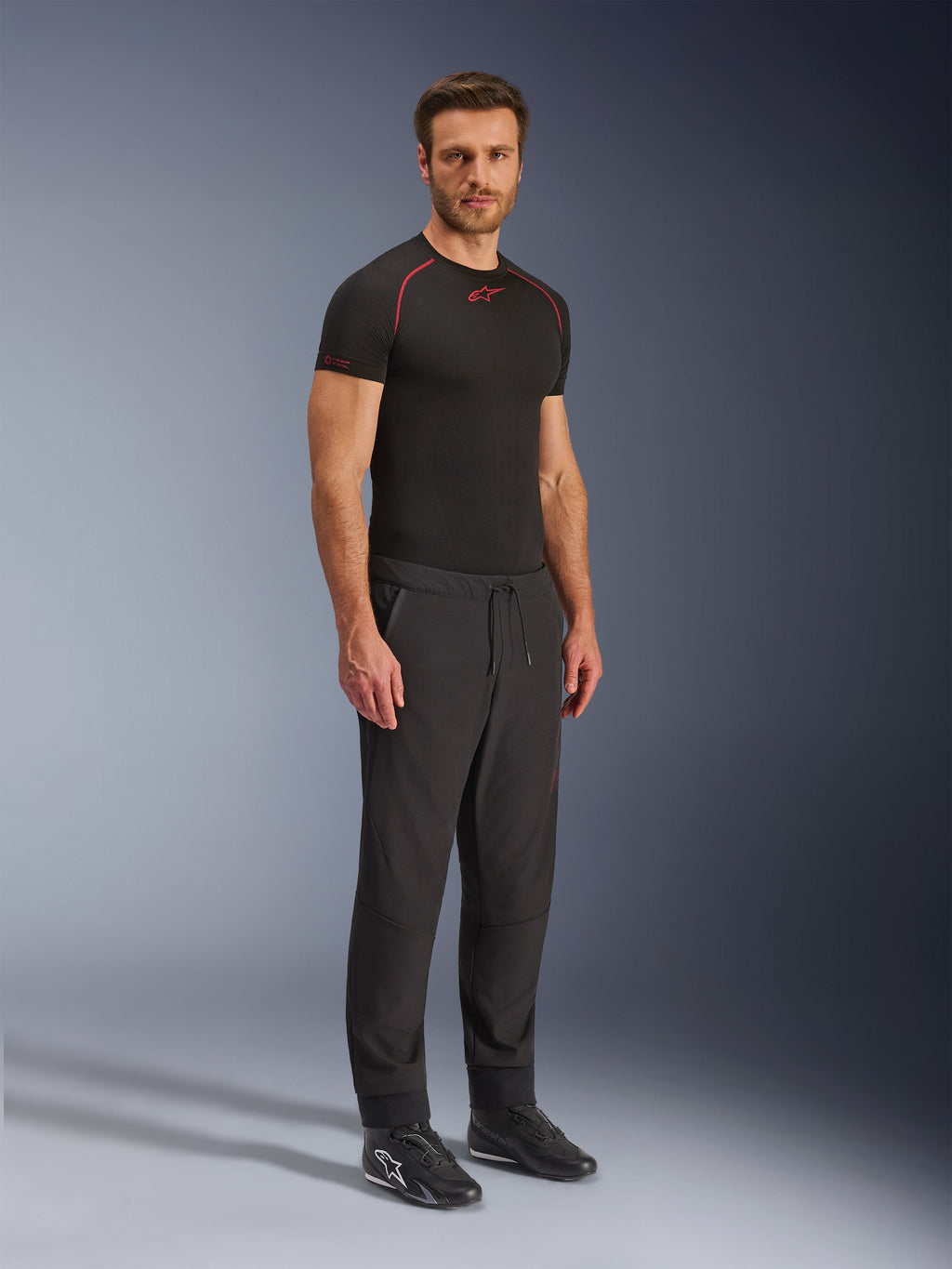 Alpinestars Aeroshell Airflow bukser, sorte motorcykelbukser, fuldkropsstudieoptagelse af en model iført softshell-bukser med snøre i taljen og elastiske manchetter, kombineret med en sort Alpinestars base layer-trøje og sorte køresko.