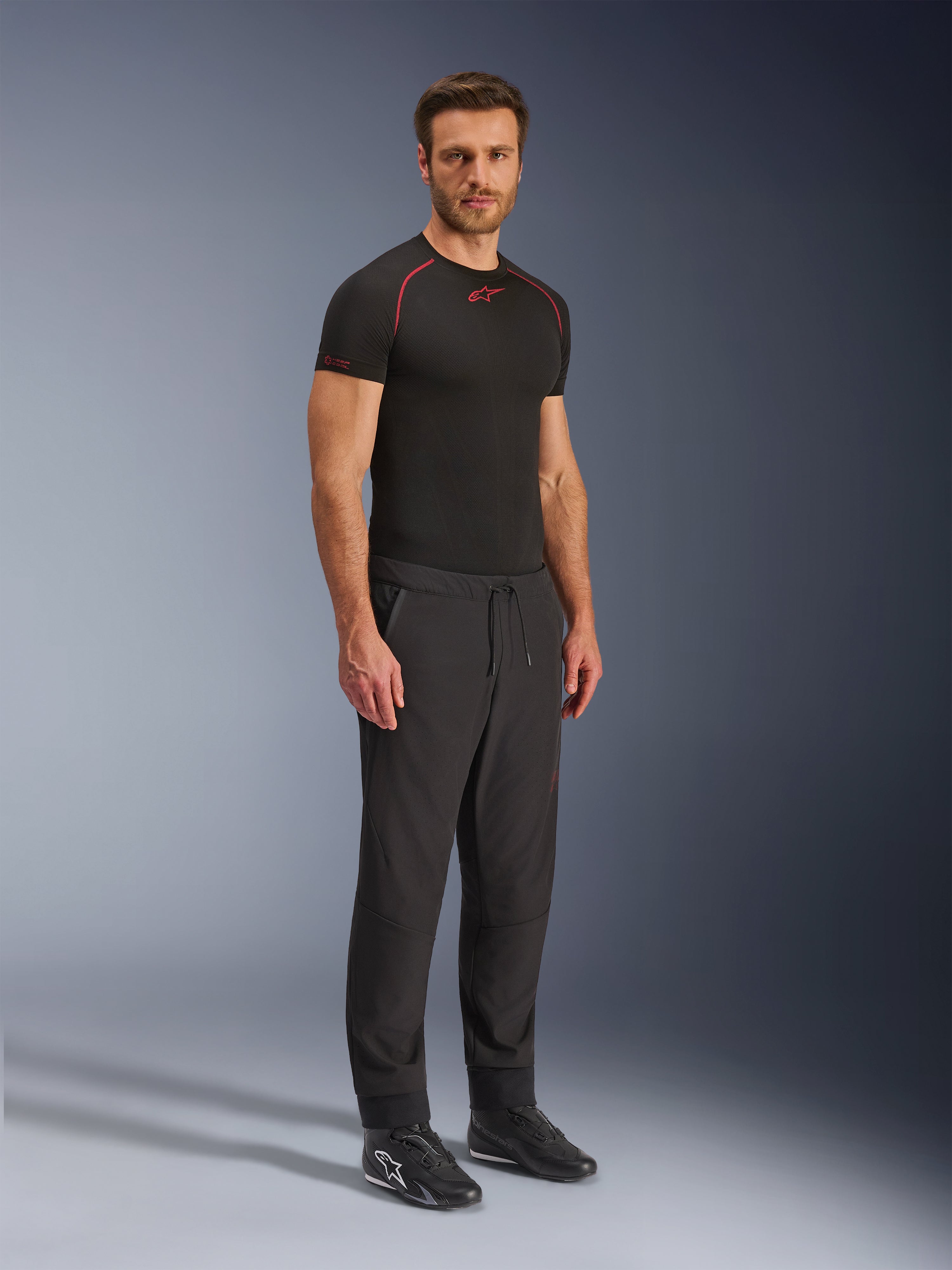 Alpinestars Aeroshell Airflow bukser, sorte motorcykelbukser, fuldkropsstudieoptagelse af en model iført softshell-bukser med snøre i taljen og elastiske manchetter, kombineret med en sort Alpinestars base layer-trøje og sorte køresko.