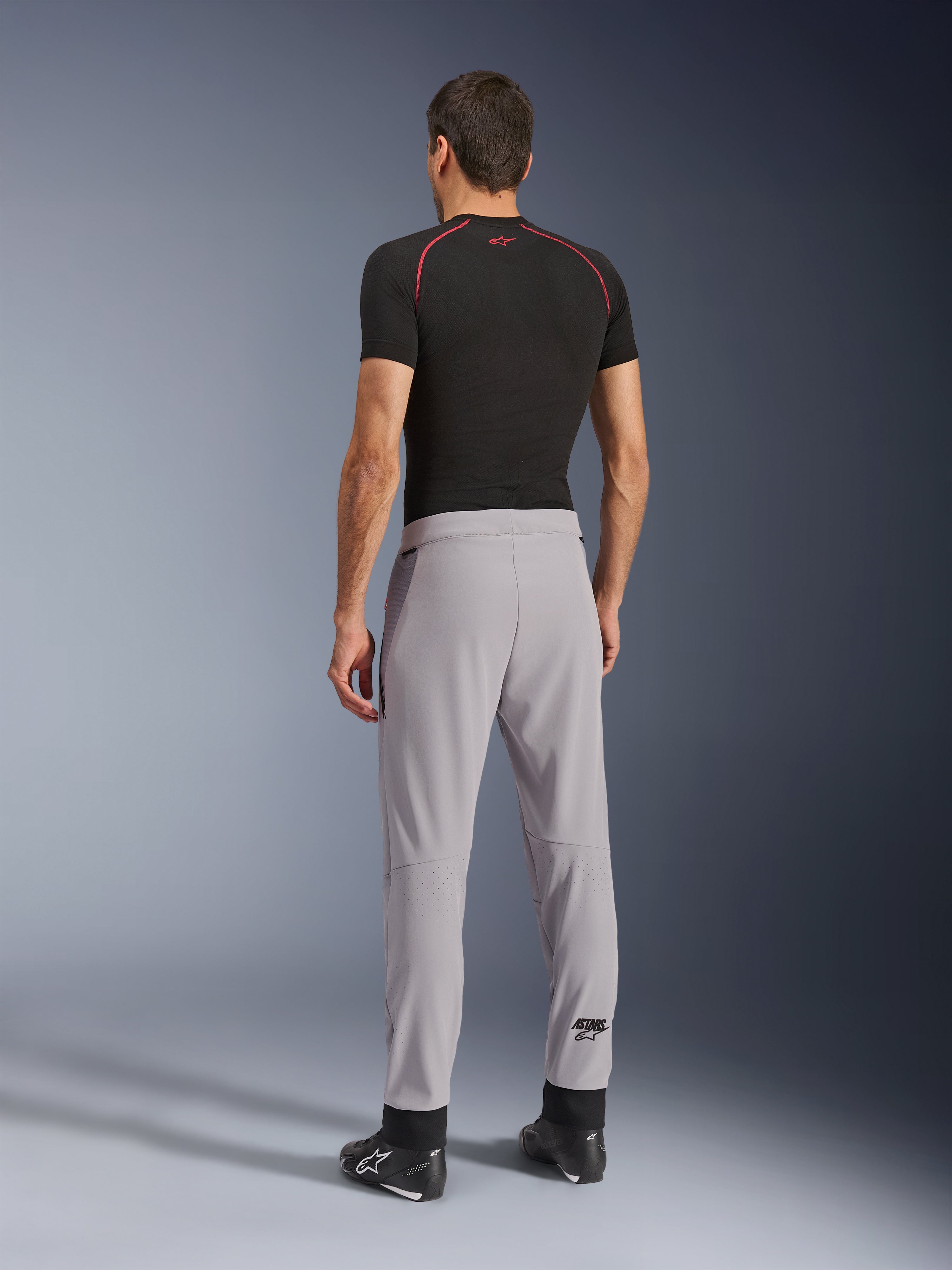 Set bagfra af en model iført Alpinestars Aeroshell Airflow bukser i Mid Gray, kombineret med en sort baselayer-trøje og sorte sko, hvilket fremhæver de laserperforerede zoner på læggene og den atletiske sportspasform.