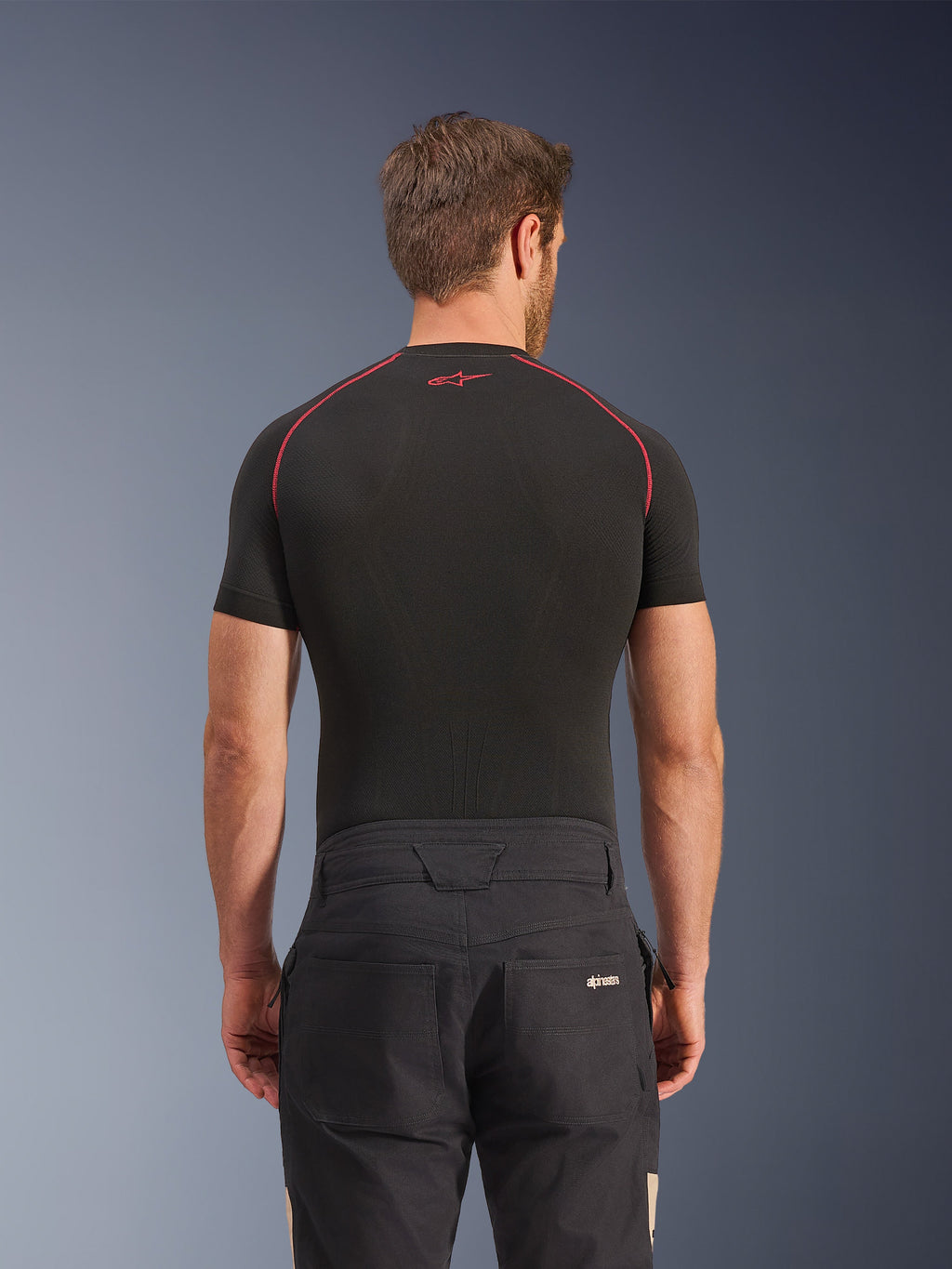 Alpinestars Flex-AST Canvas bukser i Black Aluminum, set bagfra på model, arbejdstøjsinspirerede urban motorcykelbukser med logo på baglommen, kombineret med et sort kortærmet basislag med røde syninger.