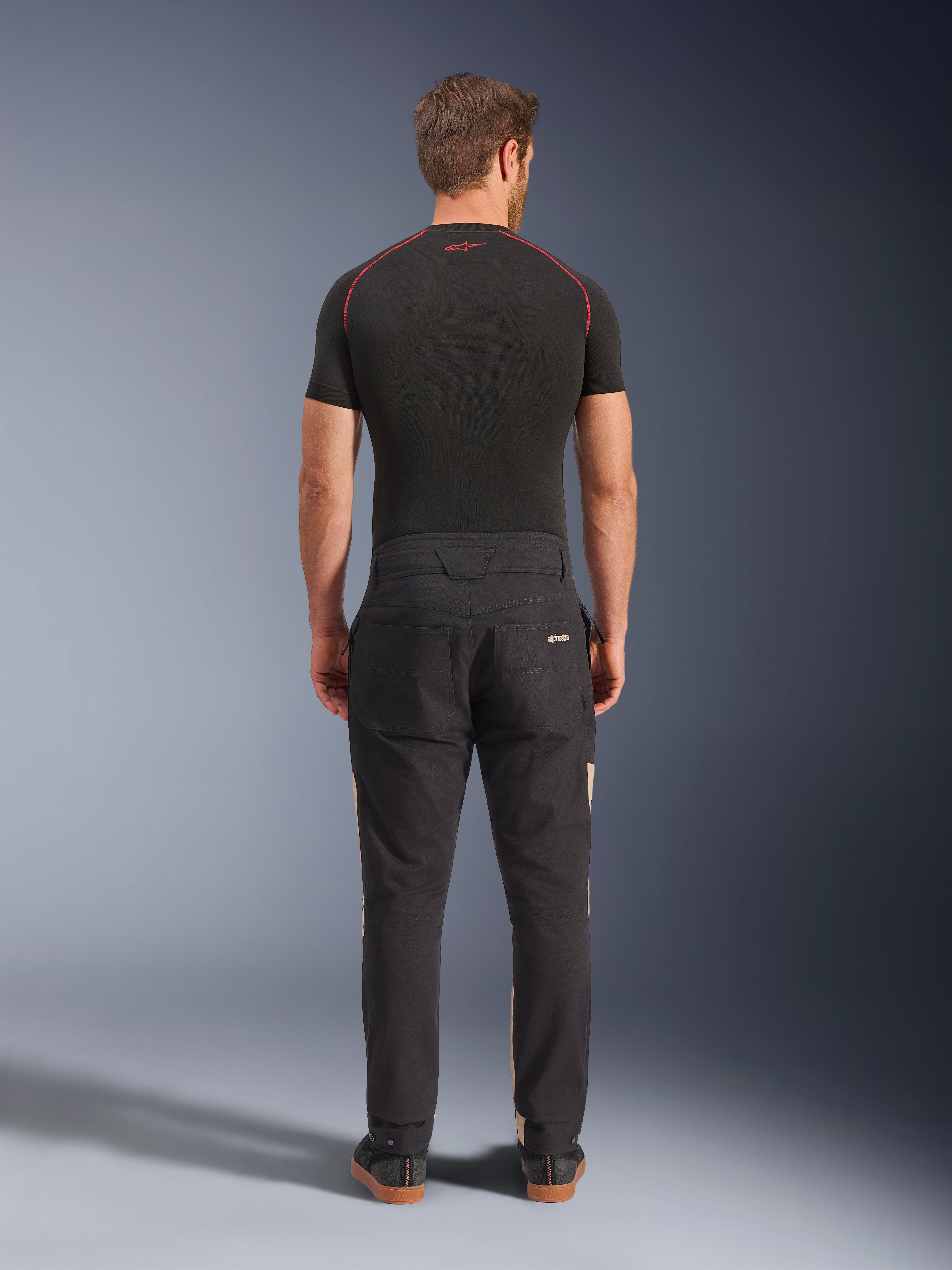 Alpinestars Flex-AST Canvas bukser i Black Aluminum, set bagfra på en model, med arbejdstøjsinspireret styling med baglommer og diskrete tan sidepaneler, båret med et sort performance-baselayer og afslappede køresneakers.