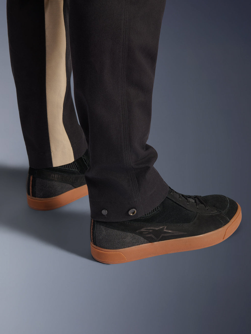 Alpinestars Flex-AST Canvas bukser i Black Aluminum, nærbillede af underbenet med trykknapjusteringer ved benåbningen og en lodret aluminiumsfarvet stribe, parret med sorte køresneakers med gummisåler, urbant motorcykeludstyr.