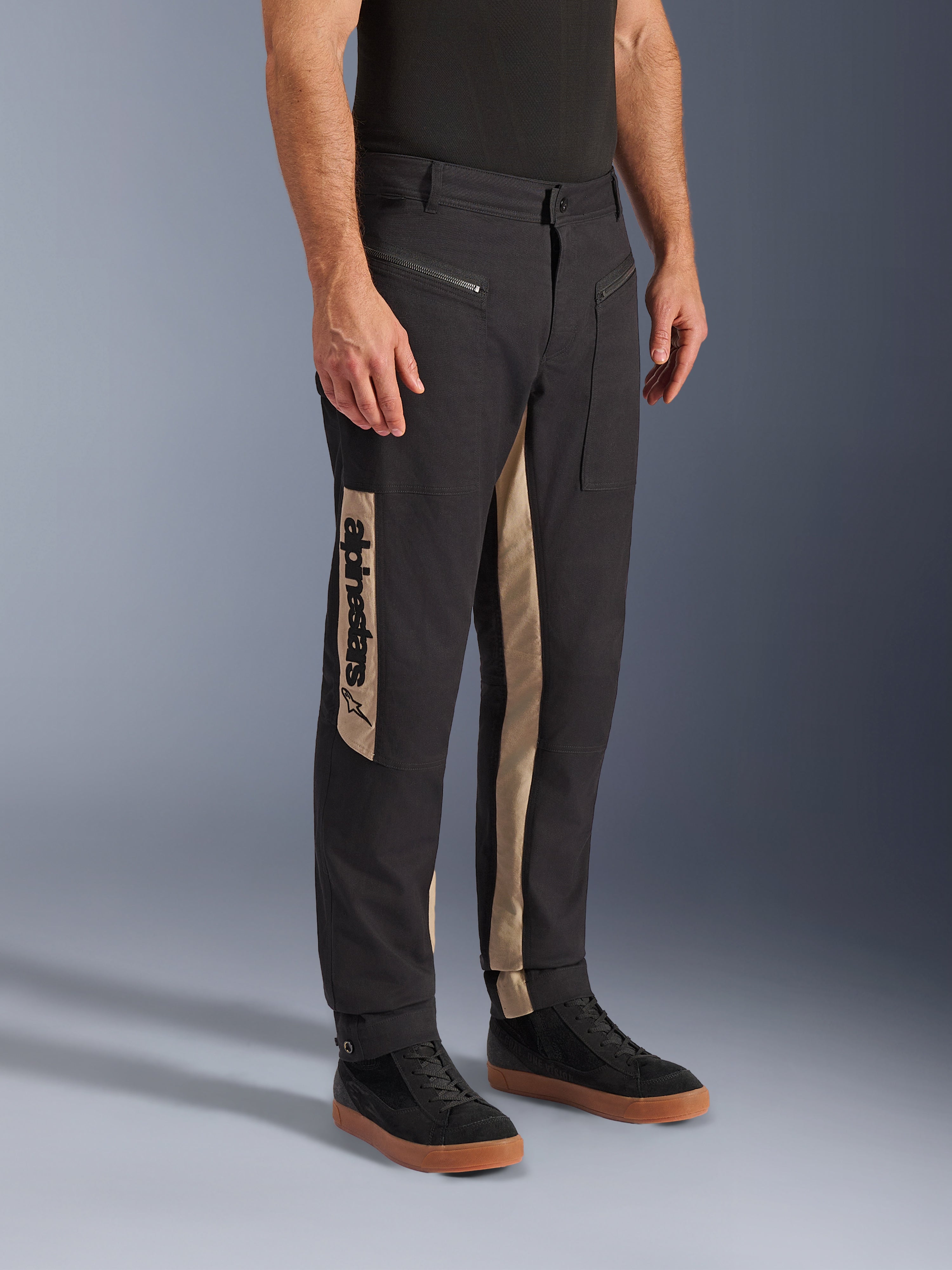 Alpinestars Flex-AST Canvas bukser, urbane motorcykelbukser, Black Aluminum, med sandfarvede sidepaneler og et lodret Alpinestars-logo, arbejdstøjsinspireret design med lynlåslommer og ergonomisk snit, båret med afslappede sorte sneakers for et moderne bylook.