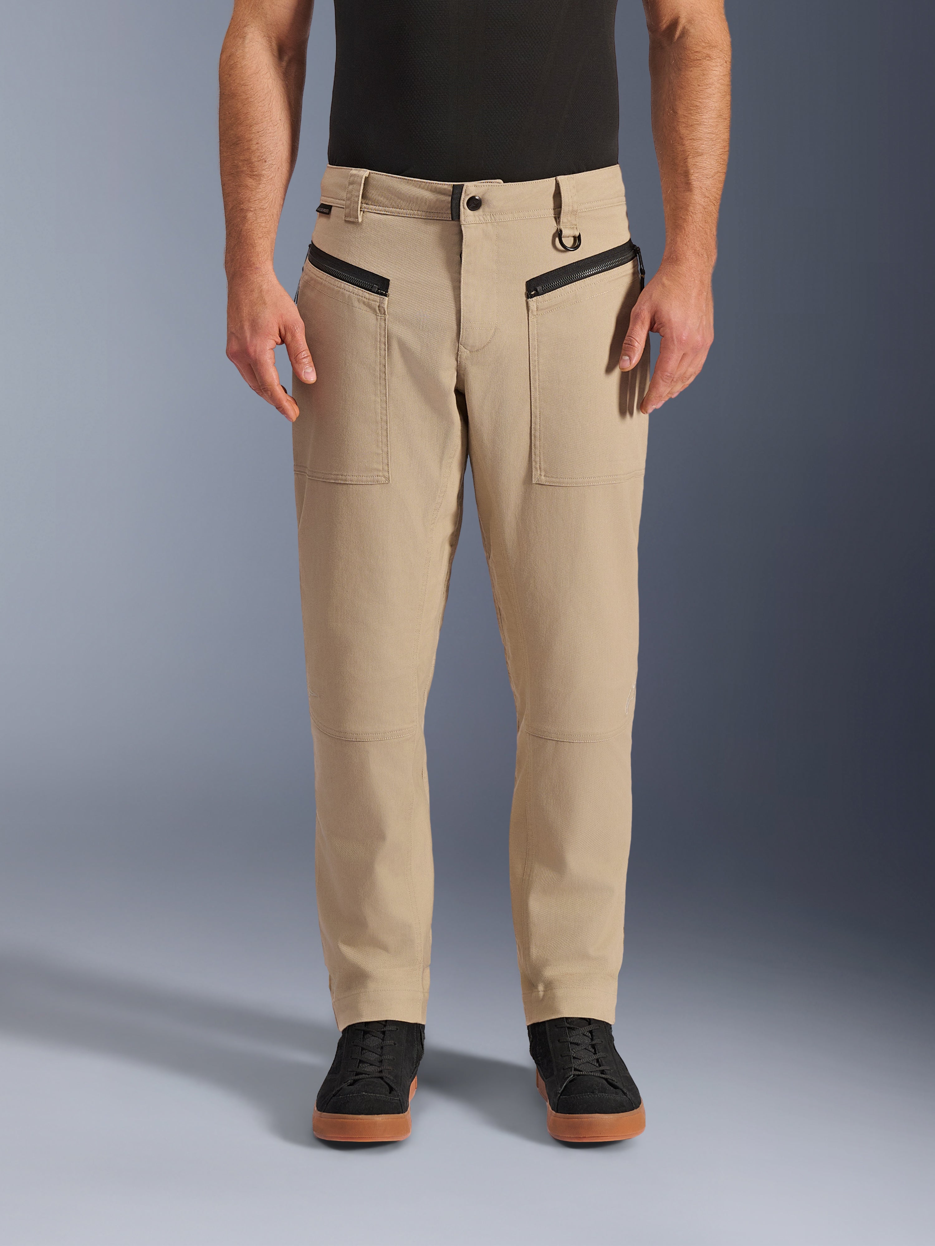 Alpinestars Flex-AST Canvas bukser, urban motorcykelbukser, brune, heritage arbejdstøjs-styling, med sorte horisontale forlommer med lynlås og D-ring i bæltestrop, båret med sort trøje og sko, studiebillede set forfra
