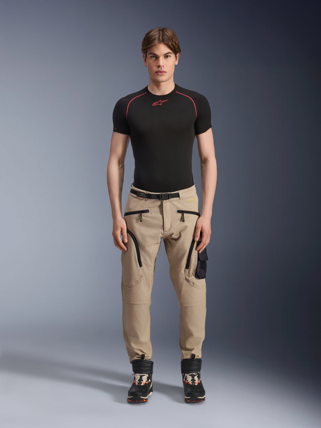Mandlig model iført Alpinestars Acteon-bukser i Sand, kombineret med et sort kortærmet basislag og sorte tekniske støvler, med cargolommer med sorte lynlåse og en tilspidset pasform, moderne urban motorcykeludstyr