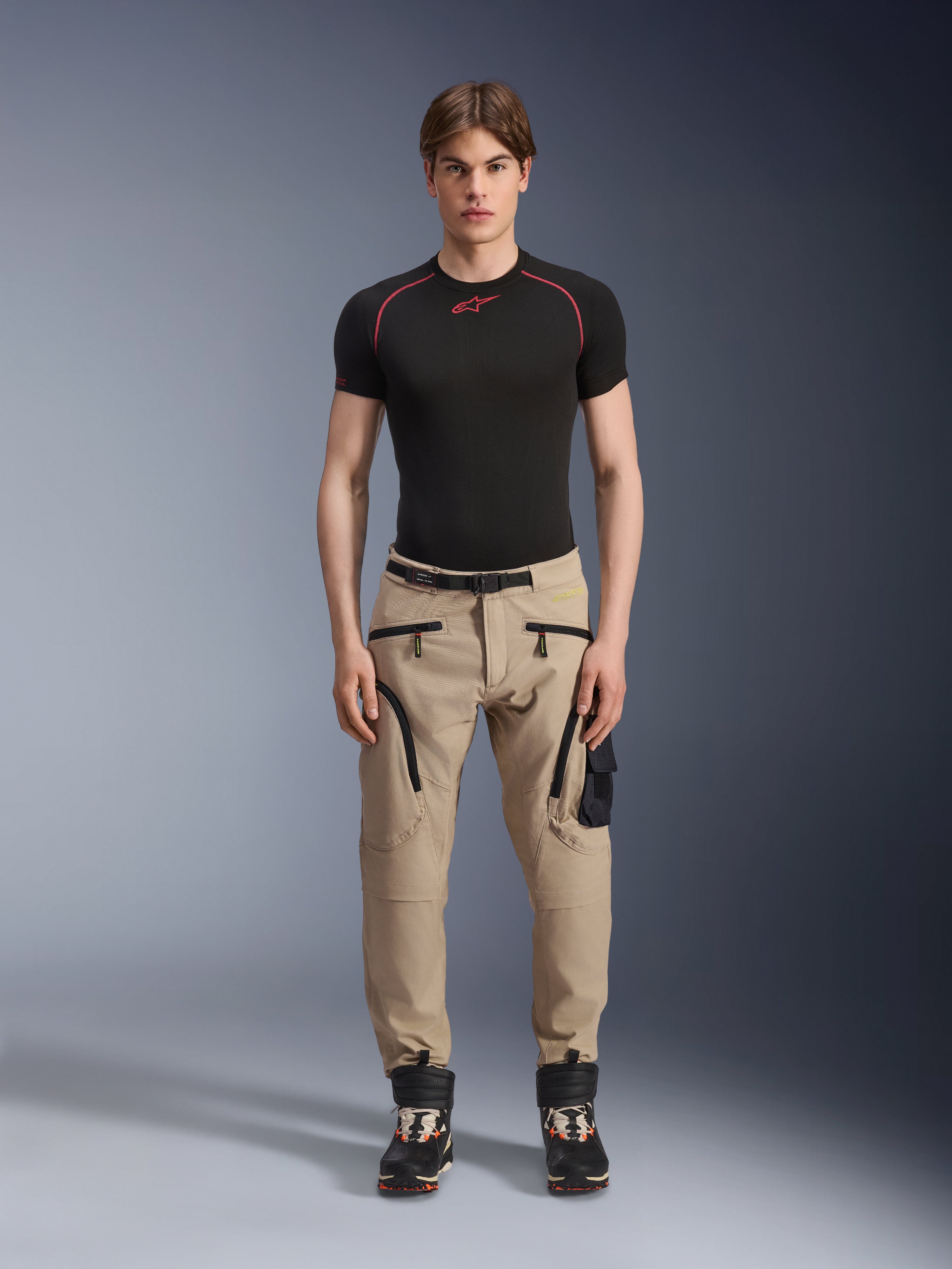 Mandlig model iført Alpinestars Acteon-bukser i Sand, kombineret med et sort kortærmet basislag og sorte tekniske støvler, med cargolommer med sorte lynlåse og en tilspidset pasform, moderne urban motorcykeludstyr