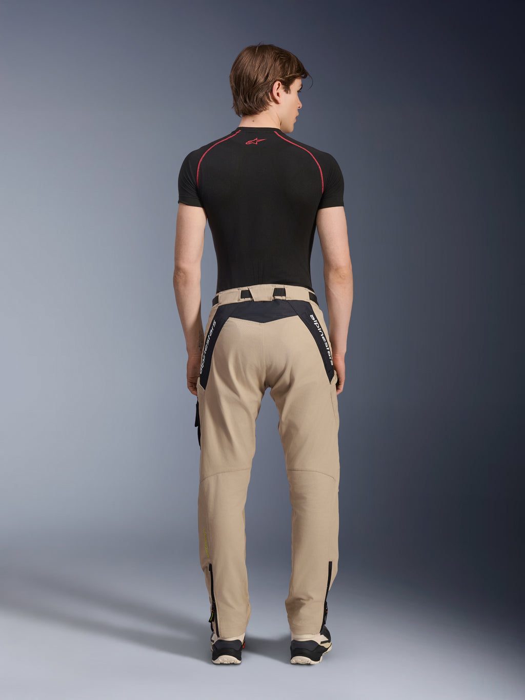 Alpinestars Acteon bukser i Sand, moderne urbane motorcykelbukser set bagfra, sorte paneler med hvid Alpinestars-branding, tilspidset pasform med lynlåse ved kanten, båret med et sort kortærmet basislag og sneakers.