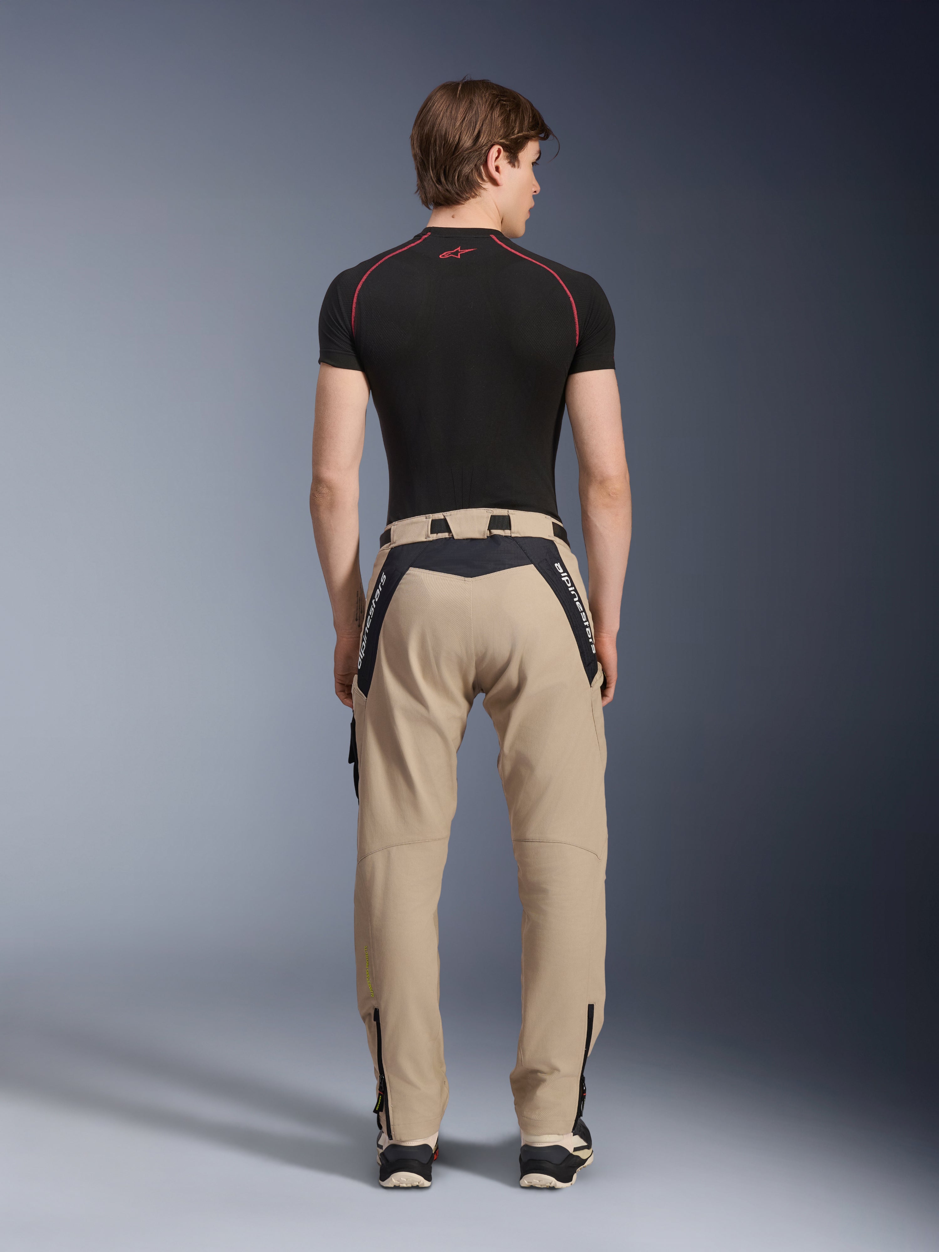 Alpinestars Acteon bukser i Sand, moderne urbane motorcykelbukser set bagfra, sorte paneler med hvid Alpinestars-branding, tilspidset pasform med lynlåse ved kanten, båret med et sort kortærmet basislag og sneakers.