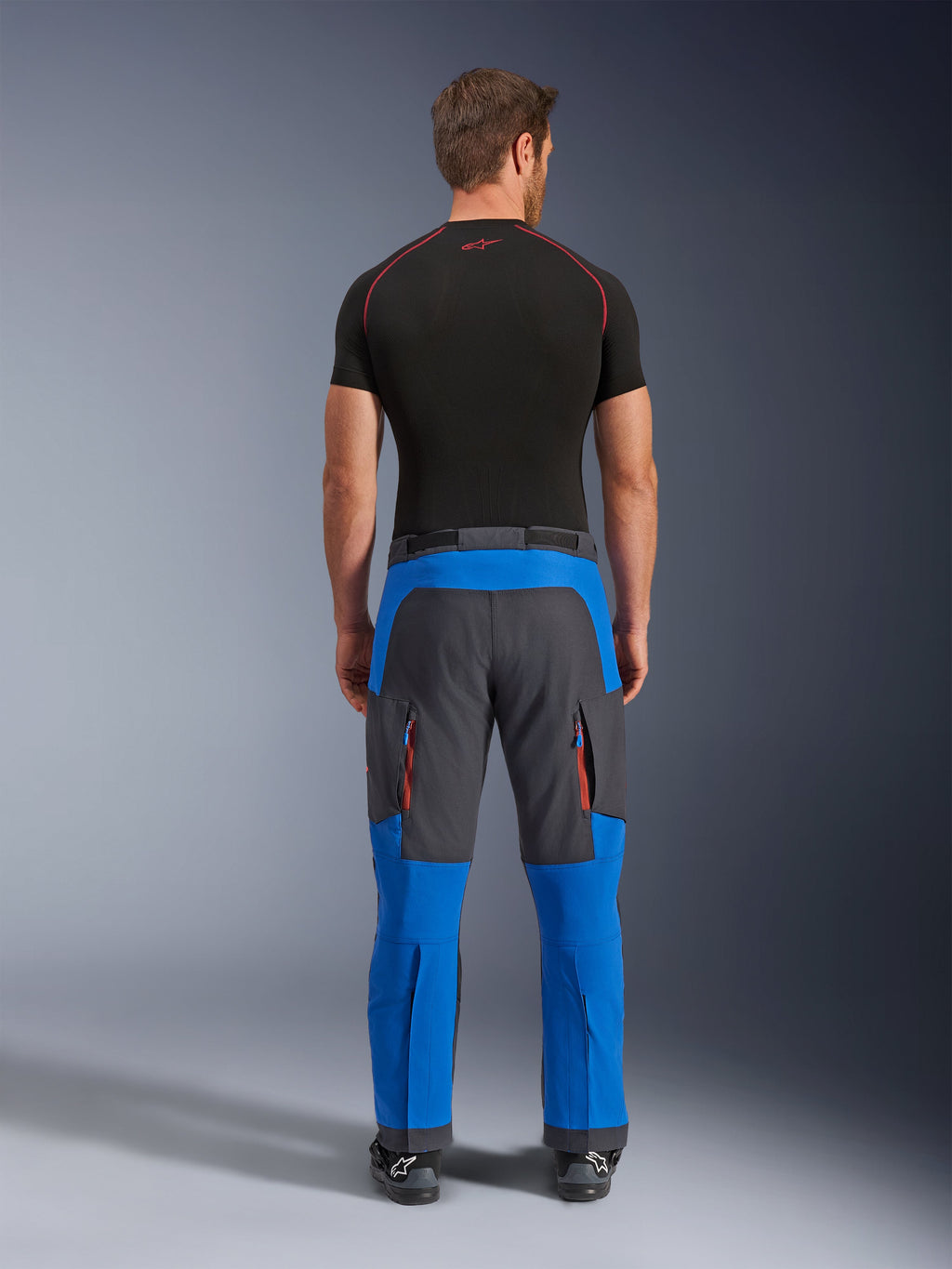Alpinestars Maxdura Dual bukser i kongeblå og mørkegrå, adventure-motorcykelbukser set bagfra, med lynlås-cargolommer med orange detaljer og et over-støvle-design, båret med et sort kortærmet basislag