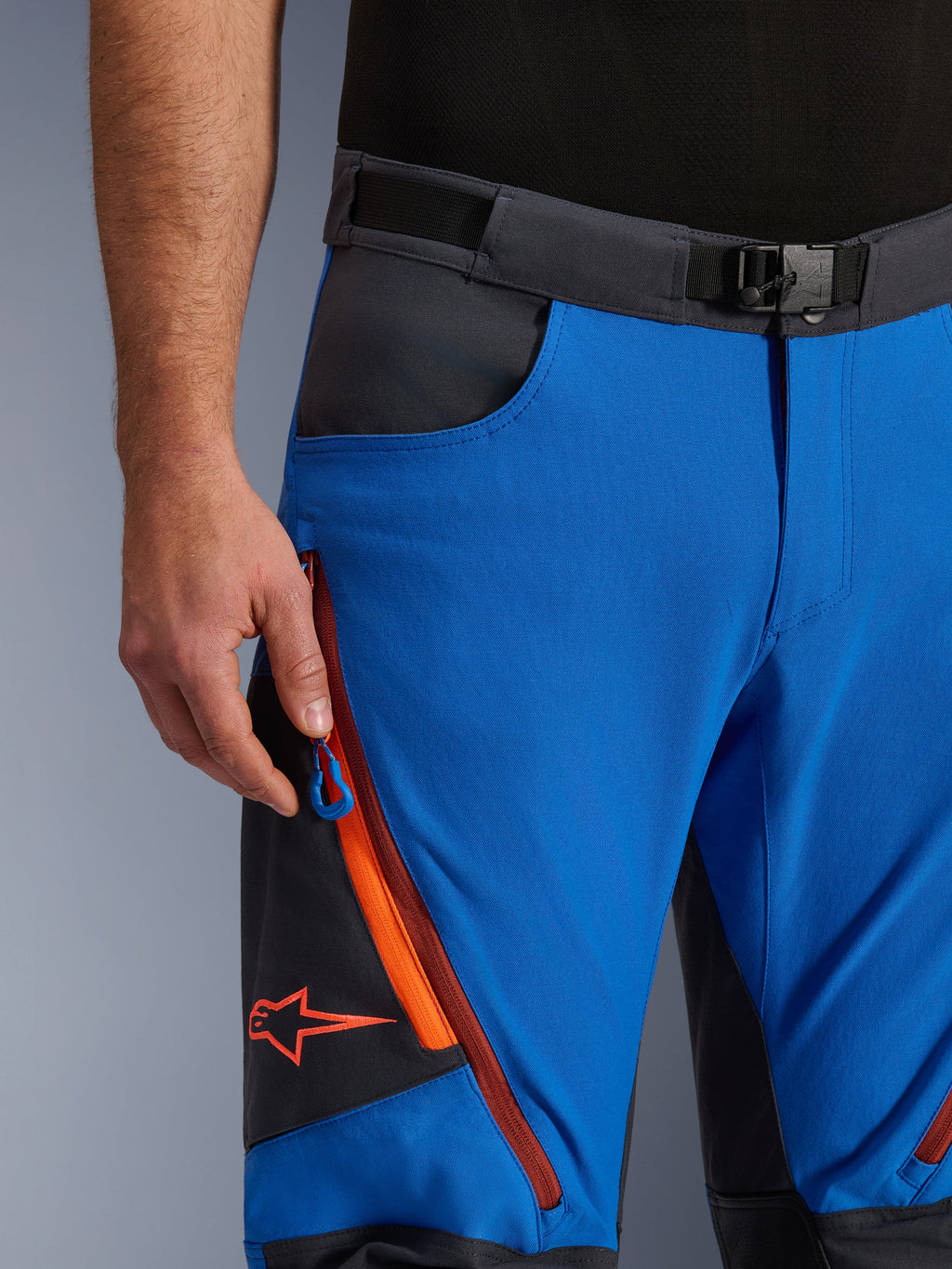 Alpinestars Maxdura Dual bukser i kongeblå og mørkegrå, nærbillede af linningen med magnetspænde, lynlåslomme med orange detaljer og rødt Alpinestars-logo, teknisk adventure-motorcykeludstyr