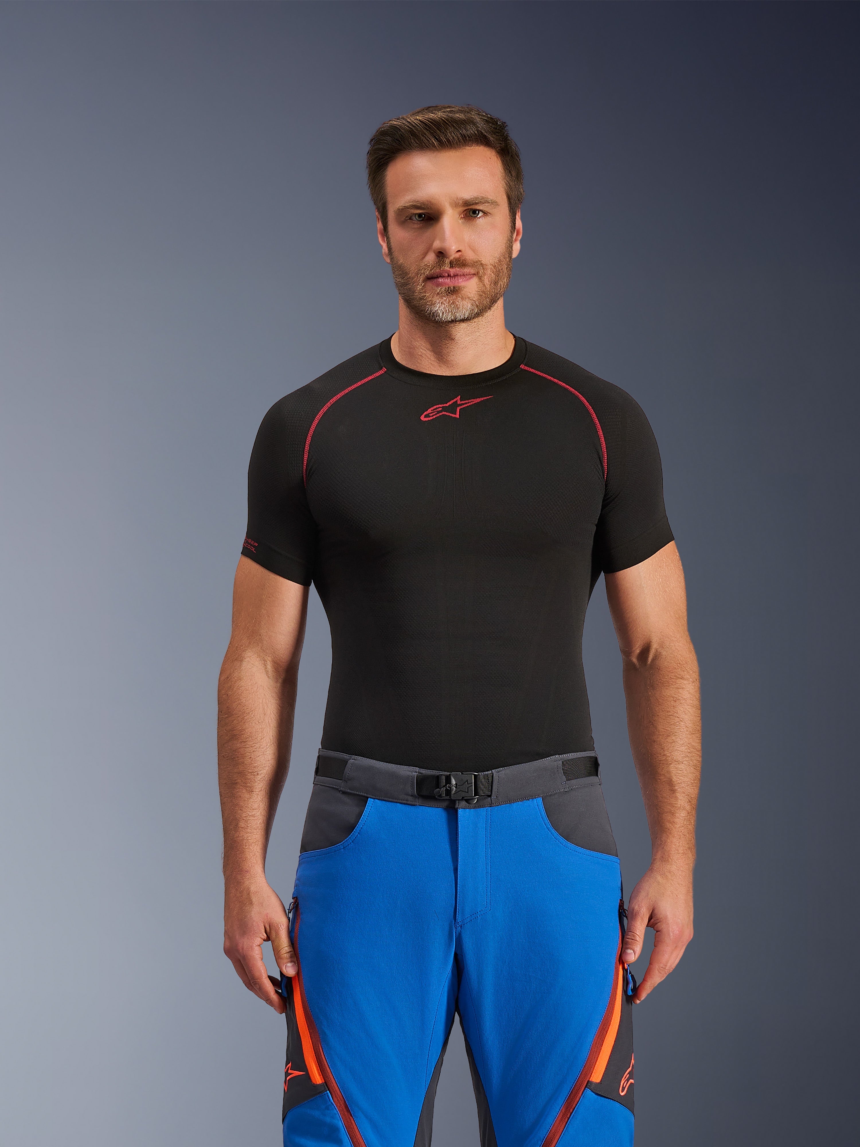 Alpinestars Maxdura Dual bukser, adventure motorcykelbukser, kongeblå og mørkegrå med orange detaljer, båret af model med sort kompressions-t-shirt, bælte med magnetspænde og lynlåsventilation på lårene, professionelt motorsportstøj