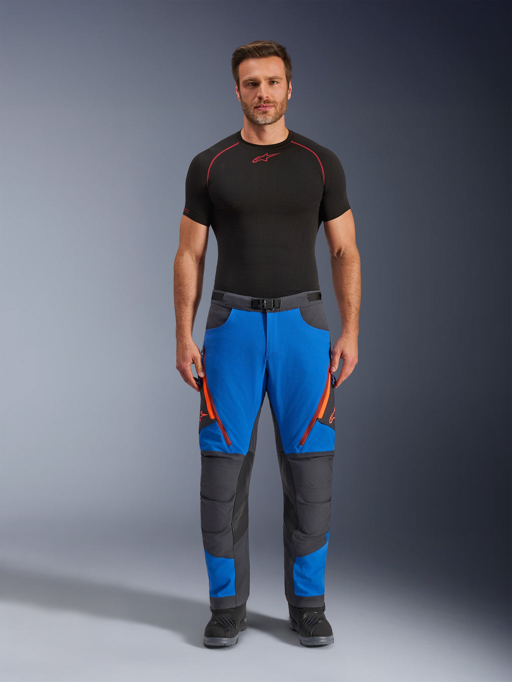 Alpinestars Maxdura Dual bukser, adventure-motorcykelbukser, kongeblå og mørkegrå, orange lynlåsdetaljer, bæres med et sort Alpinestars-basislag og sorte støvler, med forstærkede knæpaneler og et teknisk off-road-design