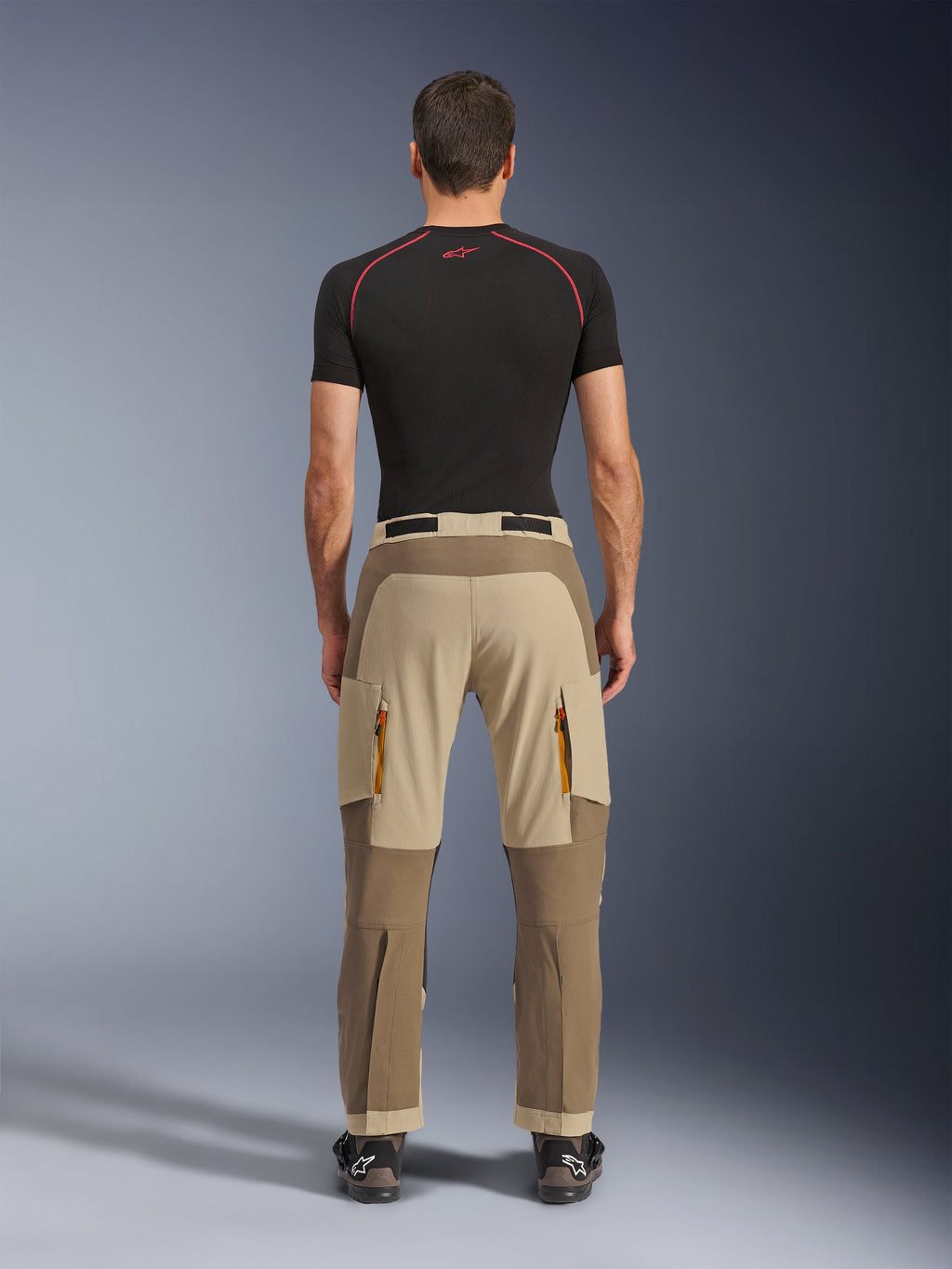 Alpinestars Maxdura Dual bukser i Walnut Aluminum, set bagfra, tofarvet brunt og beige design, adventure-kørebukser med cargolommer med lynlås og forstærket sæde, båret med en sort kompressionstrøje og motorcykelstøvler.