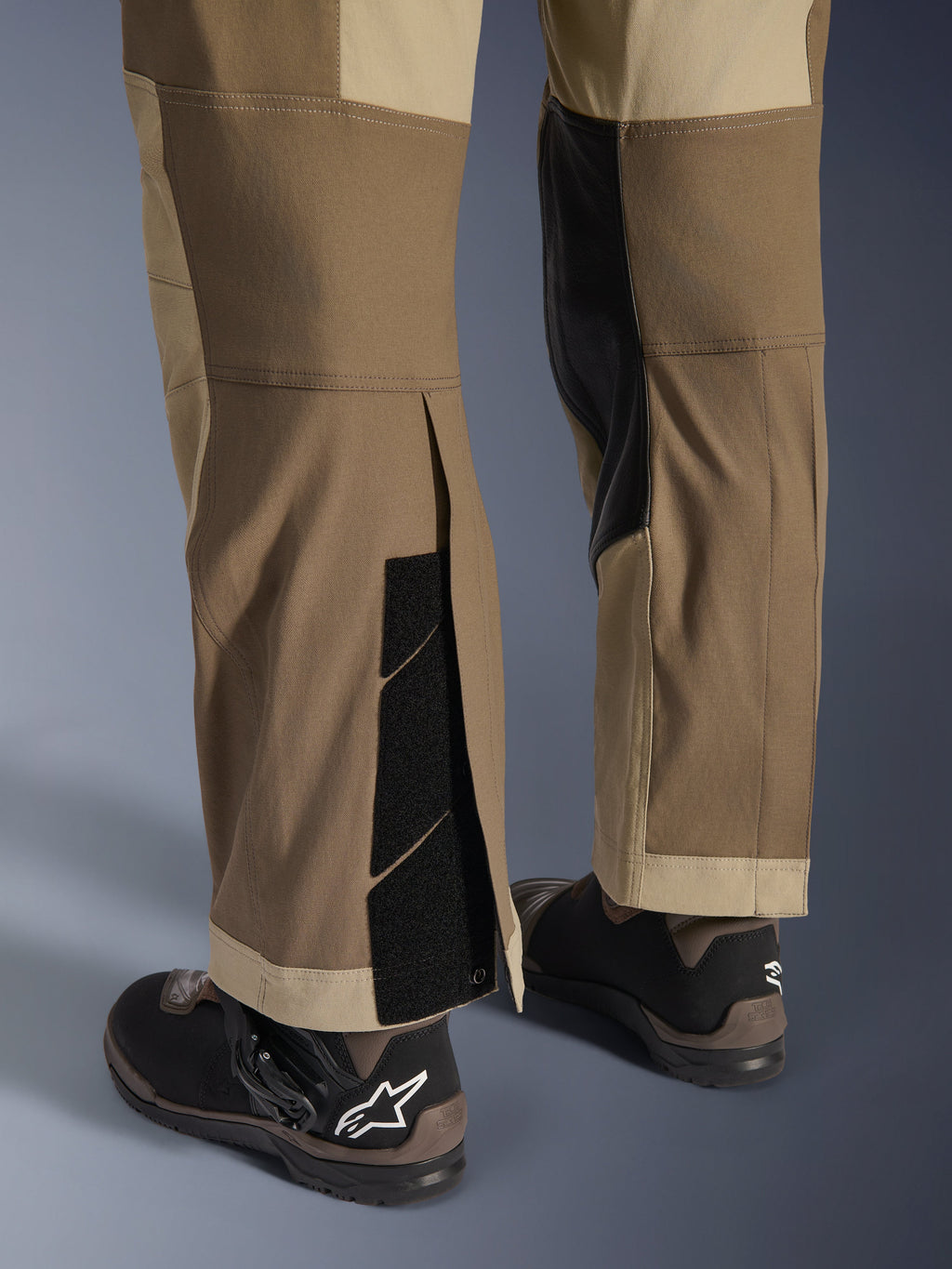 Alpinestars Maxdura Dual bukser i Walnut Aluminum, detaljeret visning af underbenet og sømmen med over-støvle-design, justerbare burrelukninger og forstærkede inderpaneler, kombineret med sorte motorcykelstøvler.