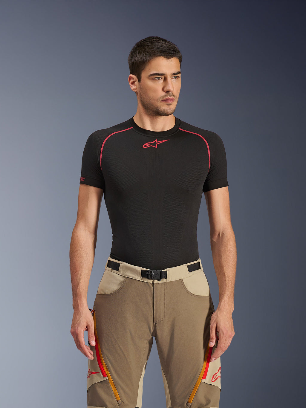 Alpinestars Maxdura Dual bukser i Walnut Aluminum, sandfarvede og beige adventure motorcykelbukser båret af en model med en sort base layer-trøje, med orange lårventilation med lynlås, rødt logo og et magnetisk taljespænde, studioportræt.