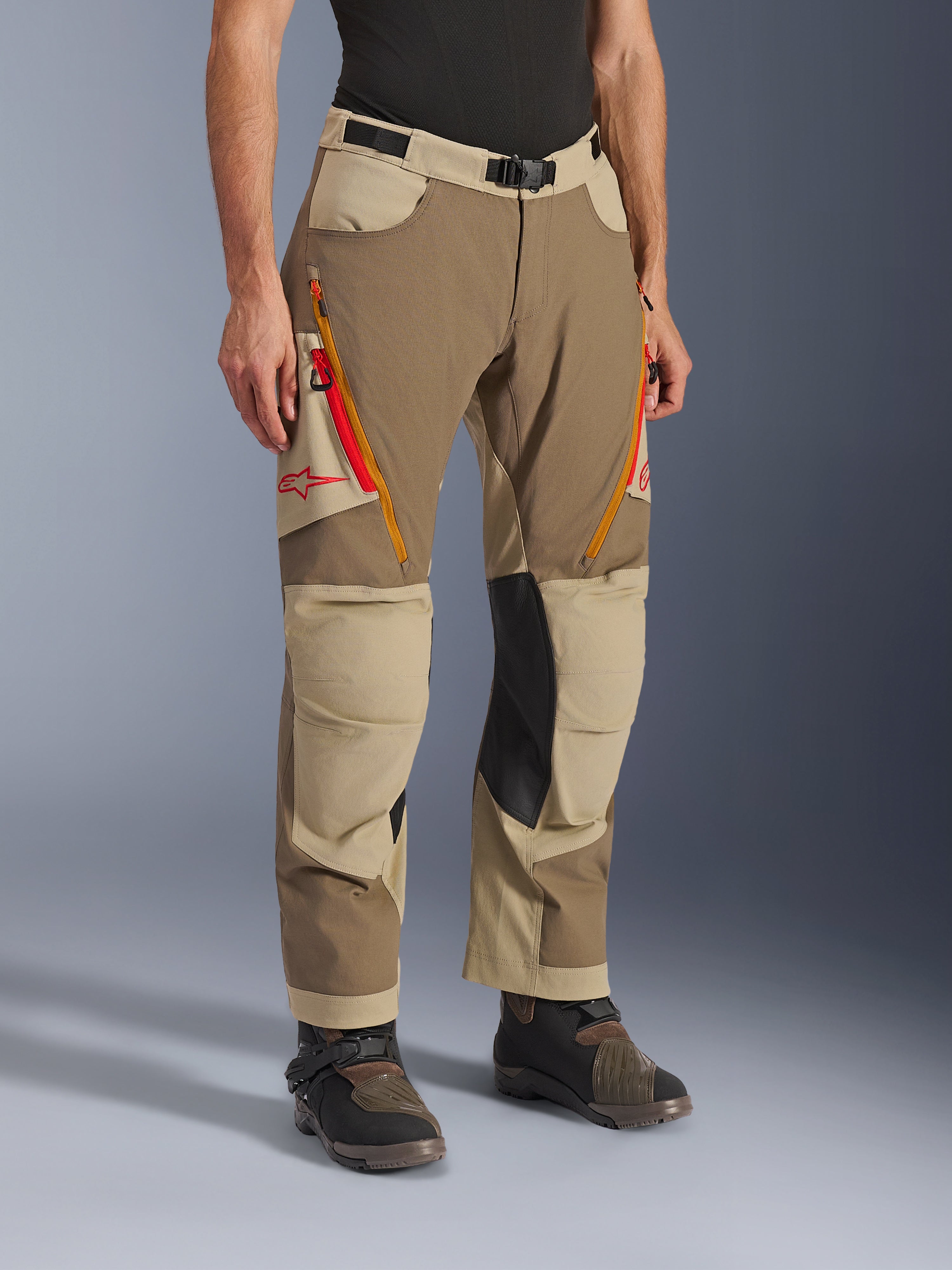 Alpinestars Maxdura Dual bukser, Walnut Aluminum brune og khaki adventure-bukser, med orange og røde ventilationsåbninger med lynlås på lårene, forstærkede inderpaneler i sort læder på knæene og rødt Astars-logo, båret med tekniske støvler.