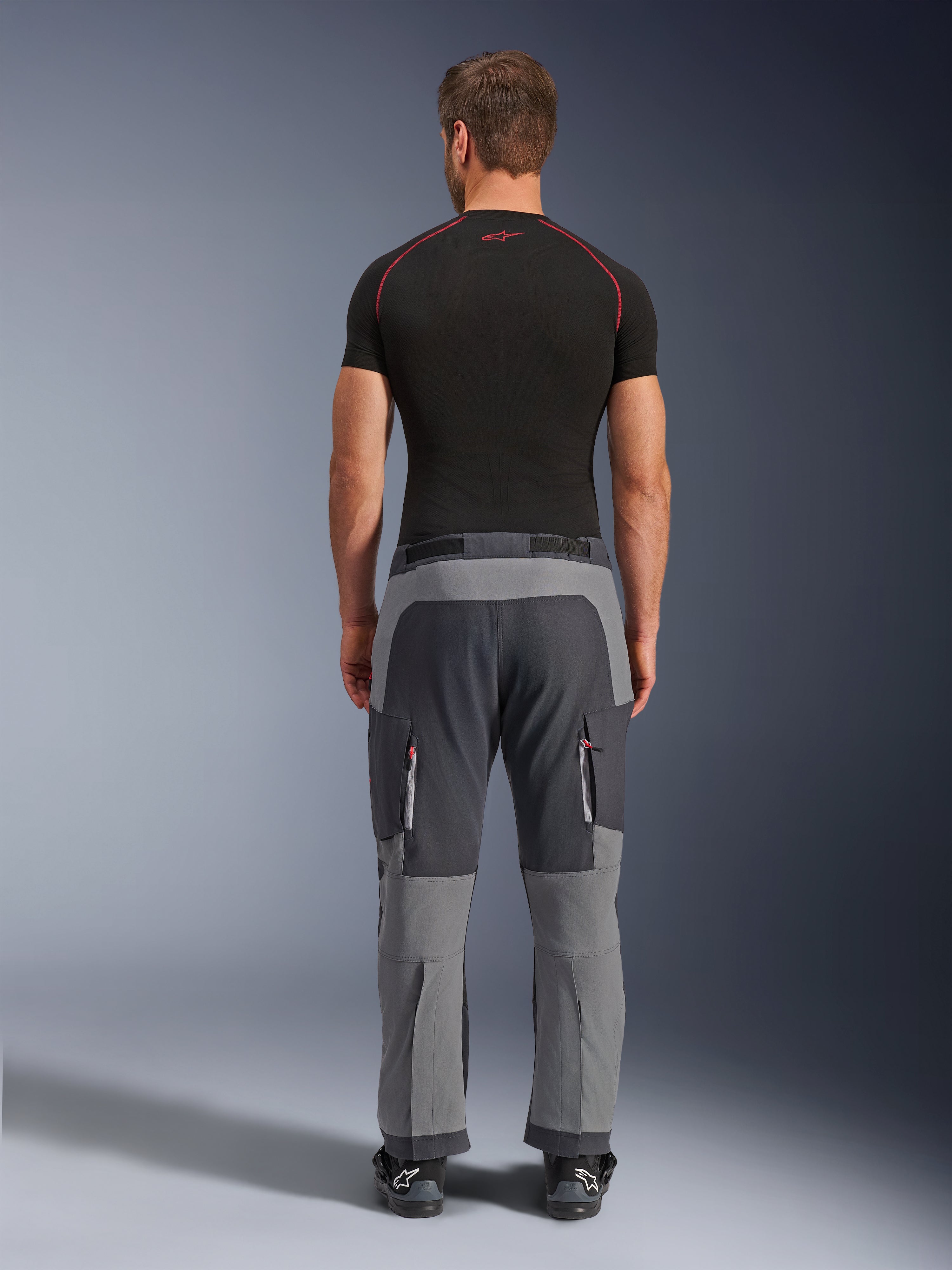 Alpinestars Maxdura Dual bukser, mørkegrå og dybgrå adventure motorcykelbukser, set bagfra på model iført sort t-shirt og støvler, med grå paneler i flere nuancer og cargolommer med lynlås og røde detaljer.